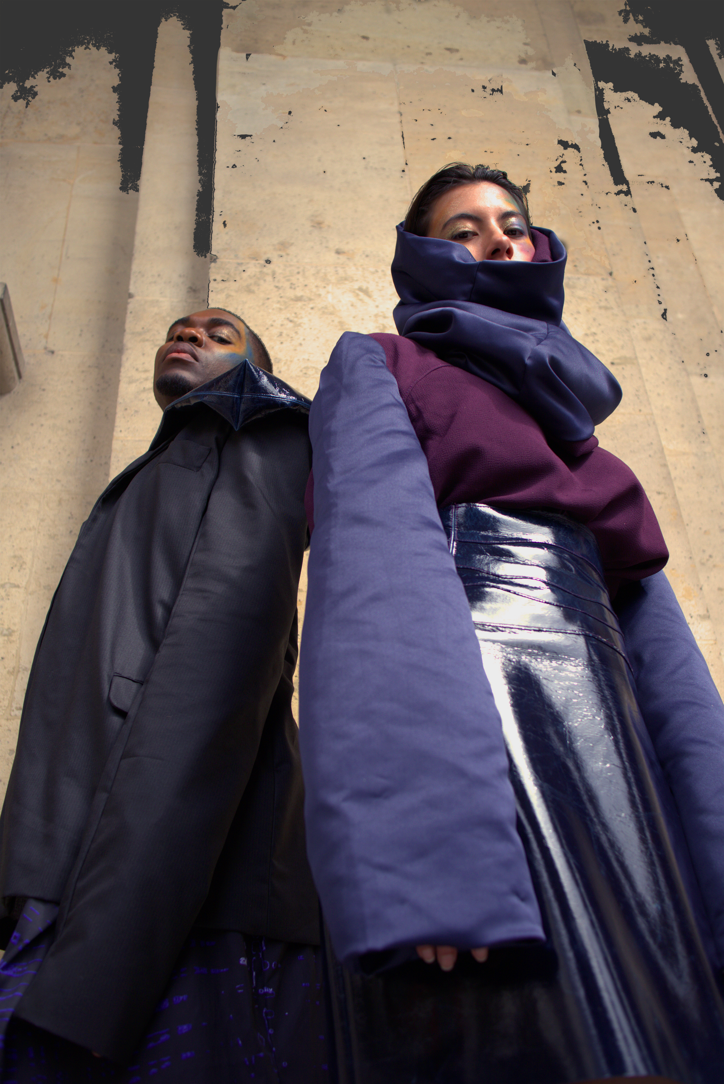 Deux personnes photographiées au sol, portant des vêtements à la mode avec des matériaux brillants et des couleurs foncées, devant un mur en pierre