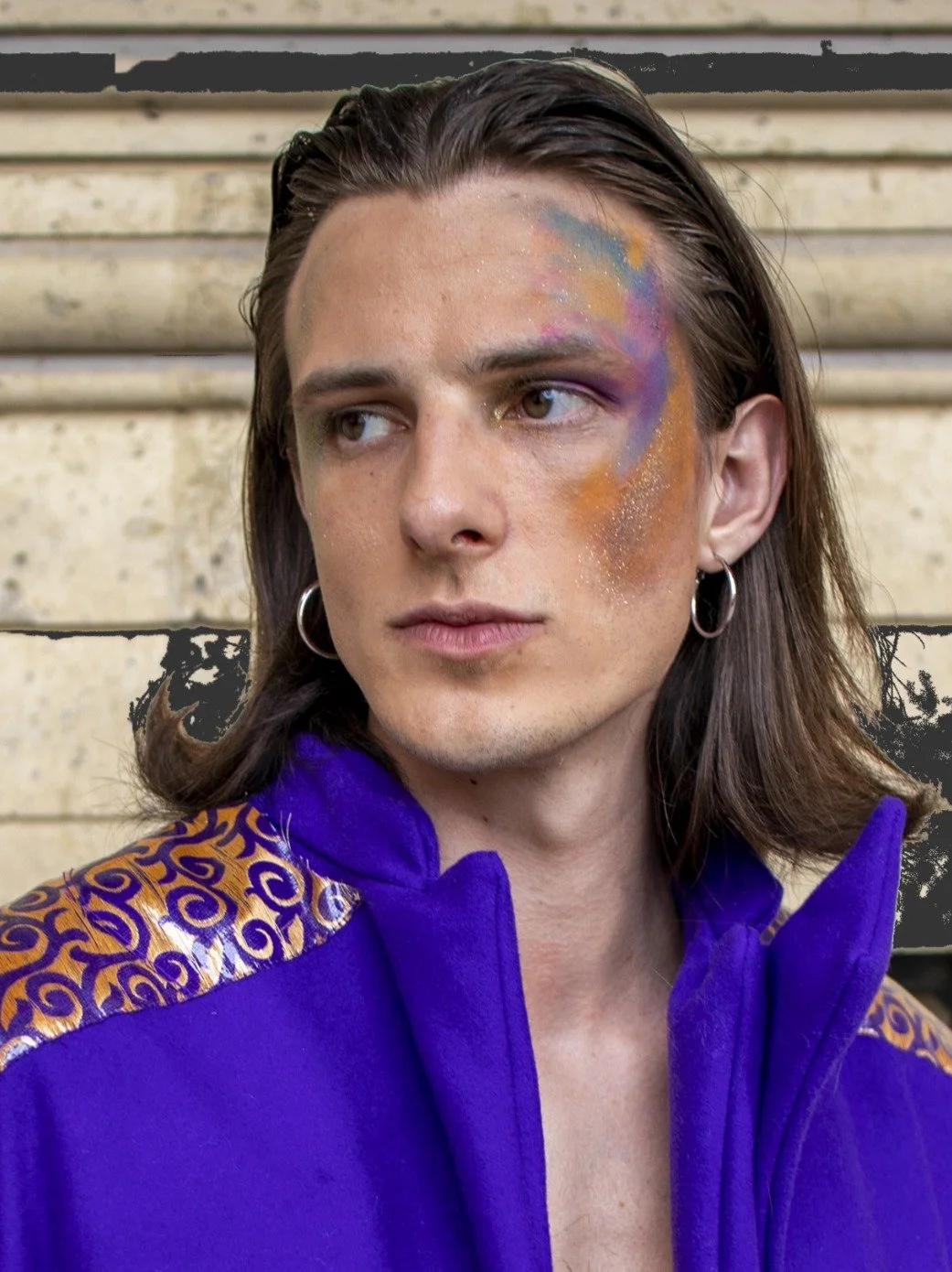 Un jeune homme avec des cheveux longs et des boucles porte un vêtement bleu avec des détails dorés sur l'épaule. Il a du maquillage coloré sur le visage, comprenant des teintes violette, orange, jaune et bleu, avec des reflets brillants. Il porte des boucles d'oreilles en cercles et regarde légèrement vers la droite.