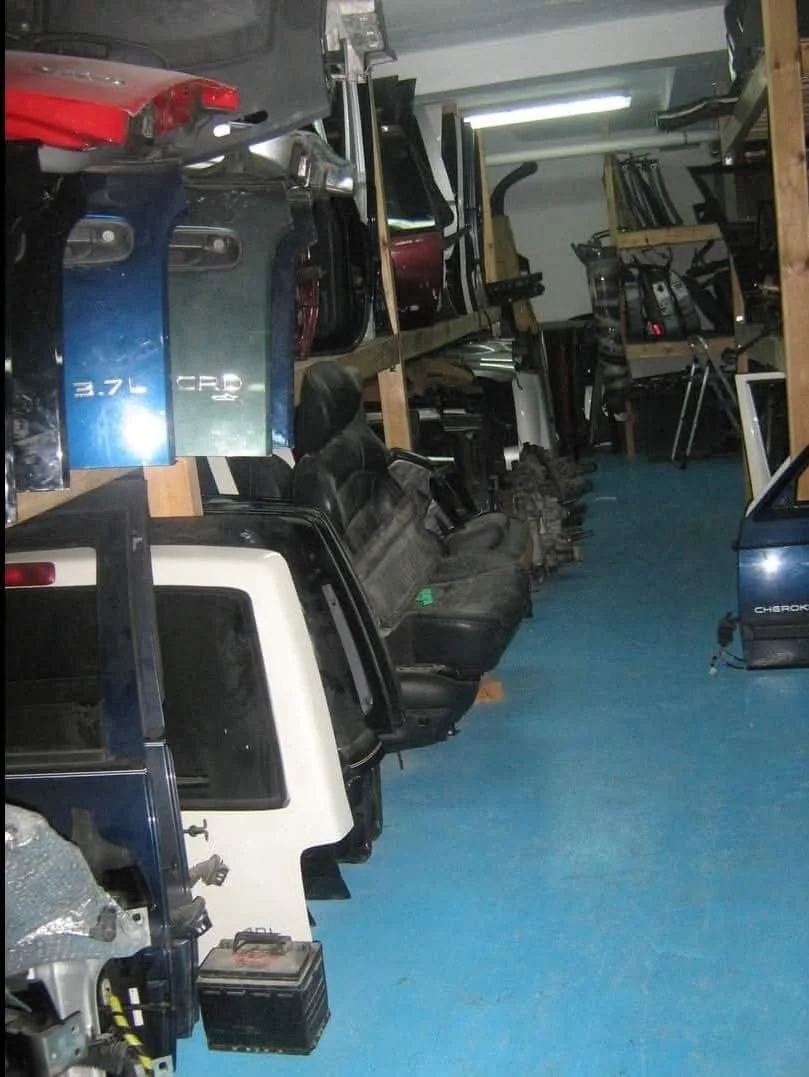 Storage room filled with car parts and seats, metal shelves holding vehicle body parts, and miscellaneous automotive tools and equipment.