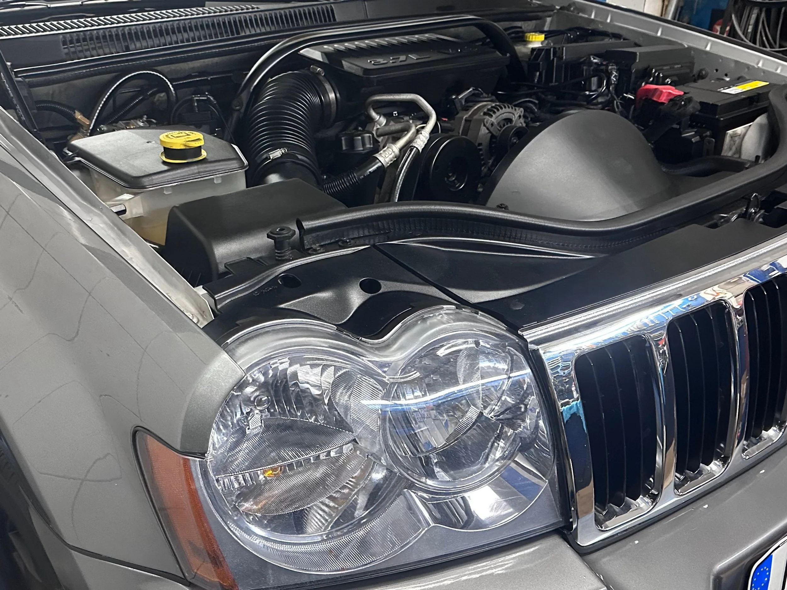 Close-up of a car engine under the hood of a silver vehicle, showing various engine components, hoses, and the headlight.