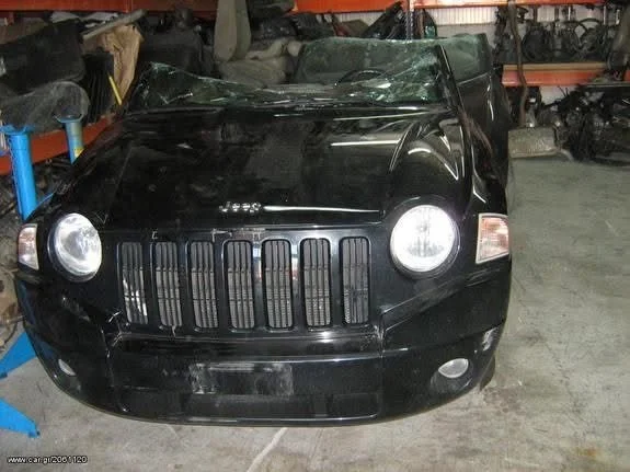 A damaged black Jeep SUV inside a warehouse or garage, with a shattered windshield and various car parts on shelves in the background.