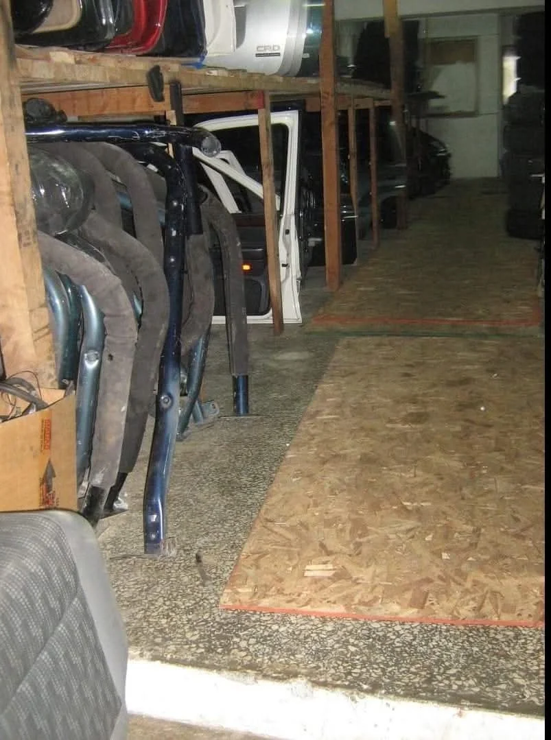 A garage or workshop with car doors and vehicle parts neatly stored on shelves and on the ground, along with wooden flooring and a partially visible seat in the foreground.