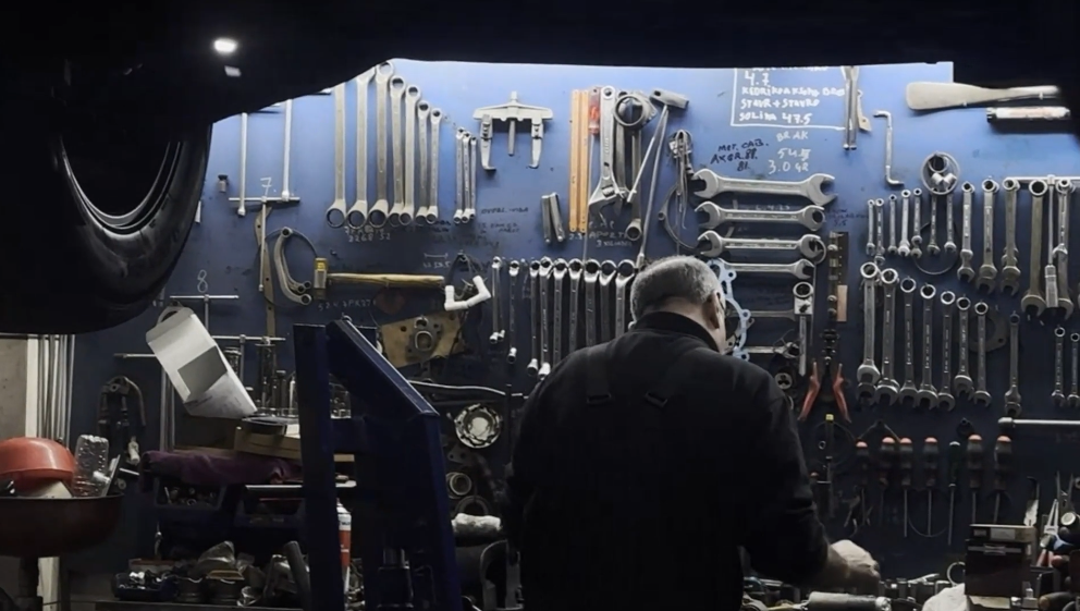 A person working at a workbench in a garage or workshop, with various tools including wrenches, pliers, and screwdrivers hanging on a blue pegboard behind them.