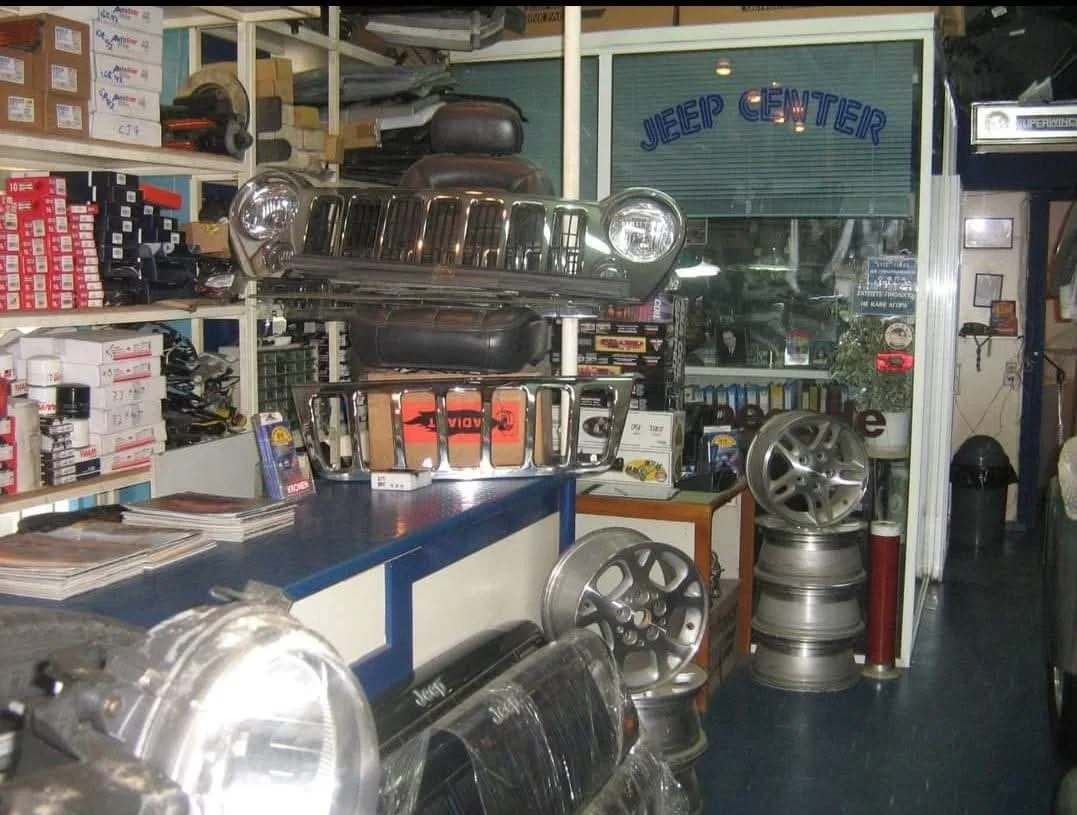 Inside an auto parts store with car wheel rims displayed, boxes and supplies on shelves, and a sign that reads 'Jeep Center' in the background.