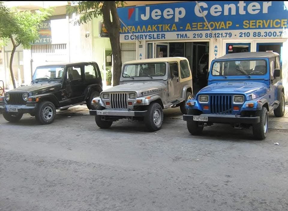 Three Jeep vehicles parked in front of a car service shop named Jeep Center, with signs in Greek and phone numbers displayed. The Jeeps include a black, a partially rusted gray, and a bright blue vehicle.