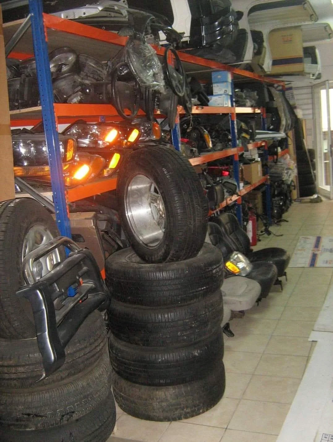Inside an auto parts store with shelves stocked with car parts, tires, and auto accessories. Several stacked tires and a shelf with vehicle lights and other automotive parts.