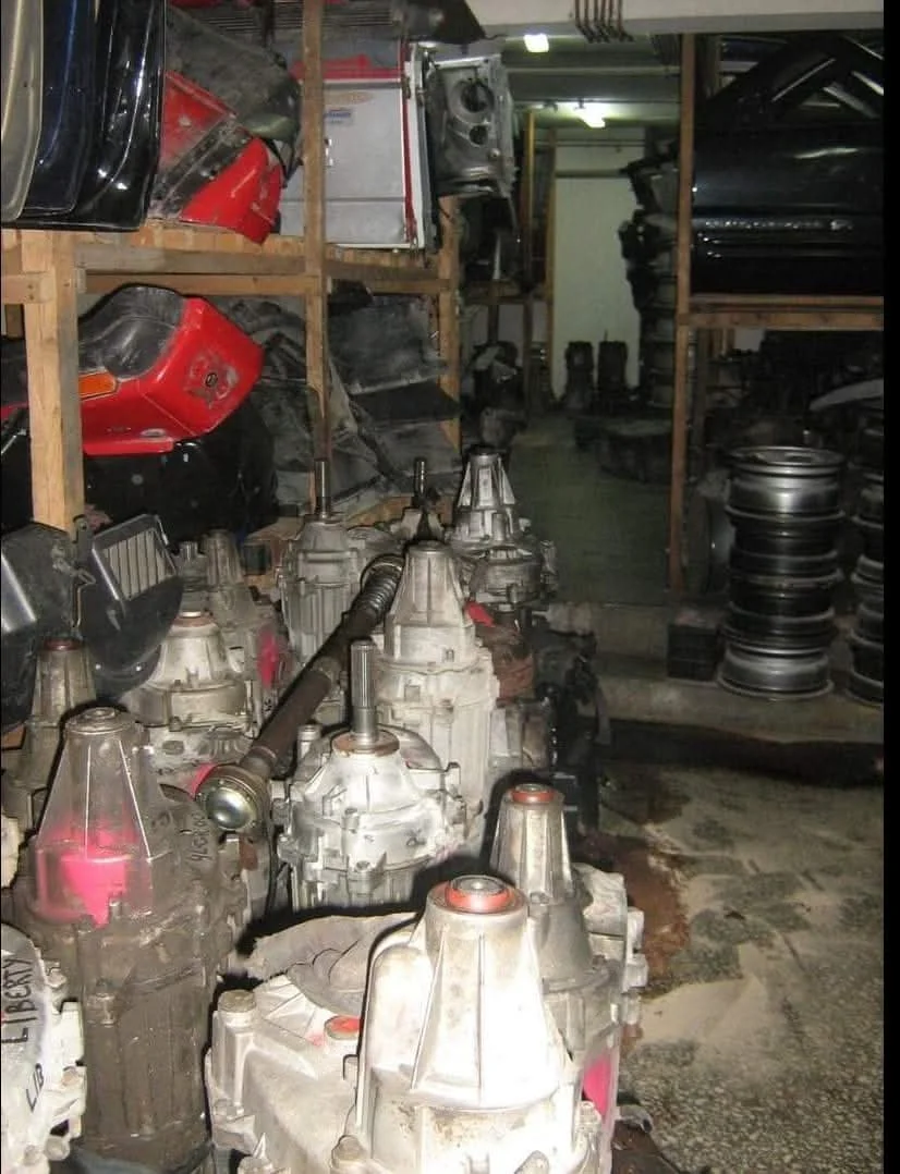 A storage room or workshop containing automotive parts, including several car driveshafts and different car bumpers stacked on shelves, with tires on the right side.