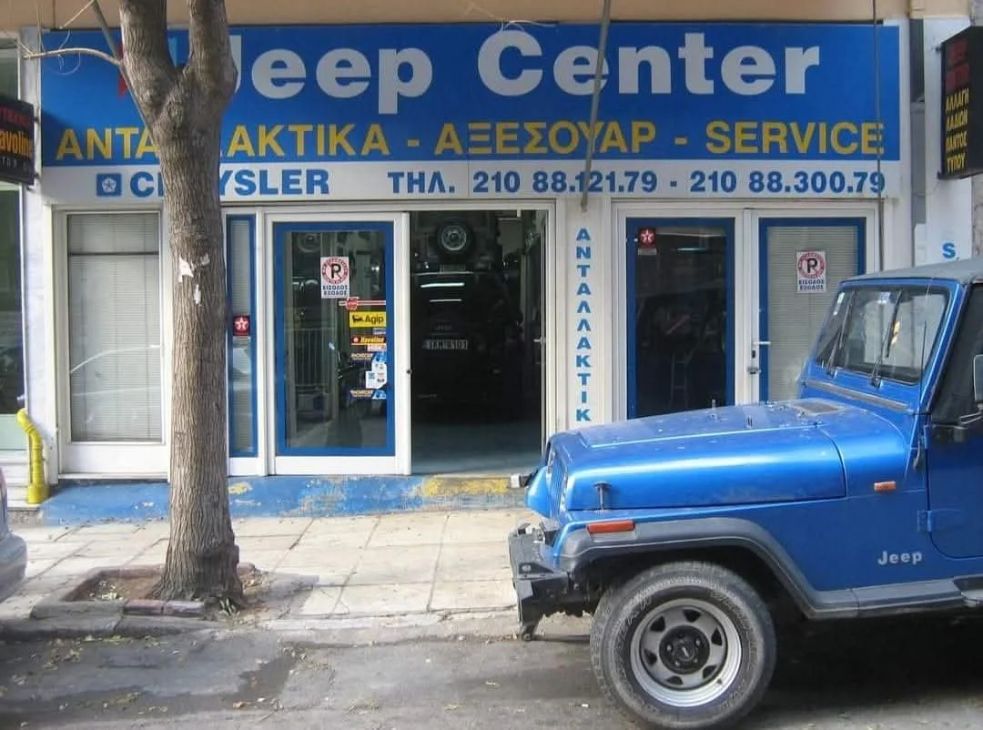 Front view of a car repair shop with a sign reading 'Jeep Center' and additional service descriptions in Greek. A blue Jeep, showing front damage, is parked outside on the street, near a tree.