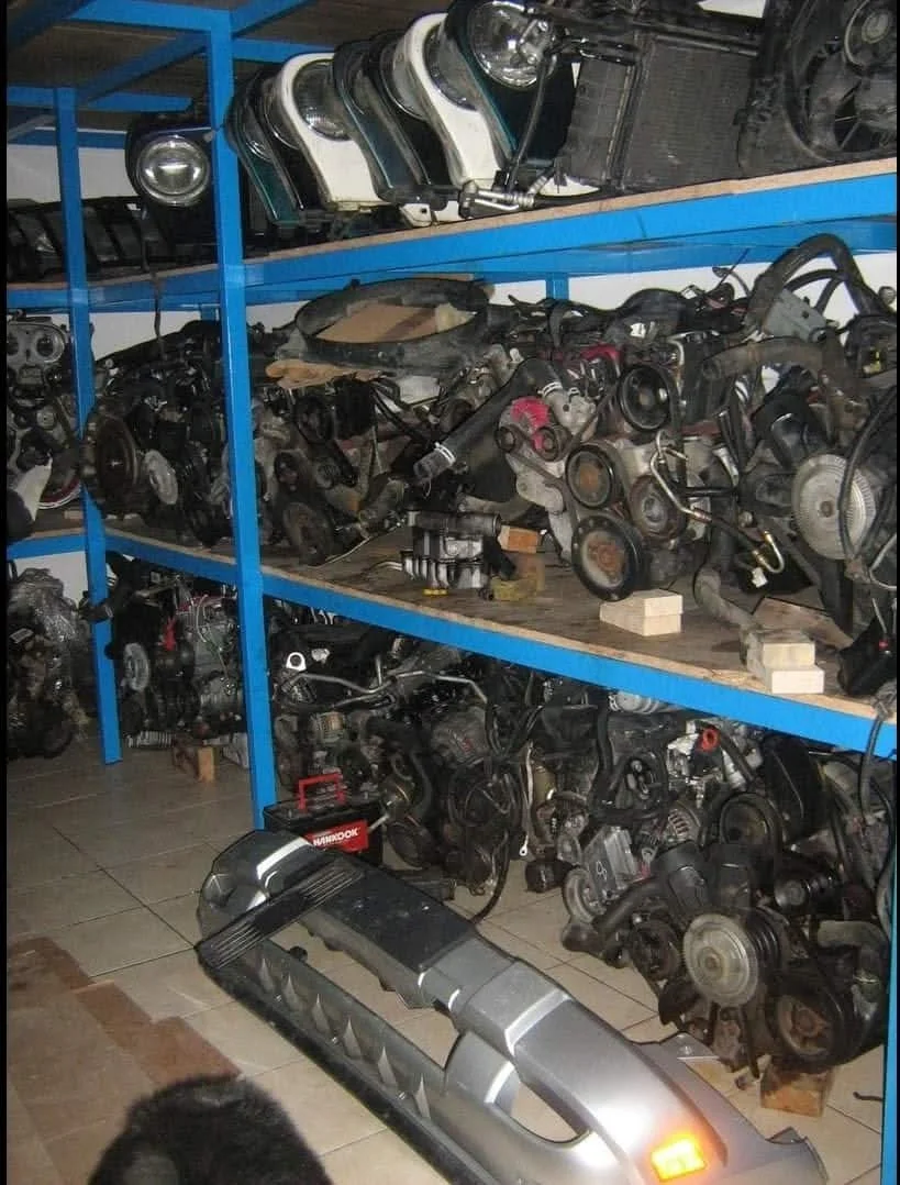 Shelves filled with car engine parts and a car bumper on the floor in a workshop or storage room.