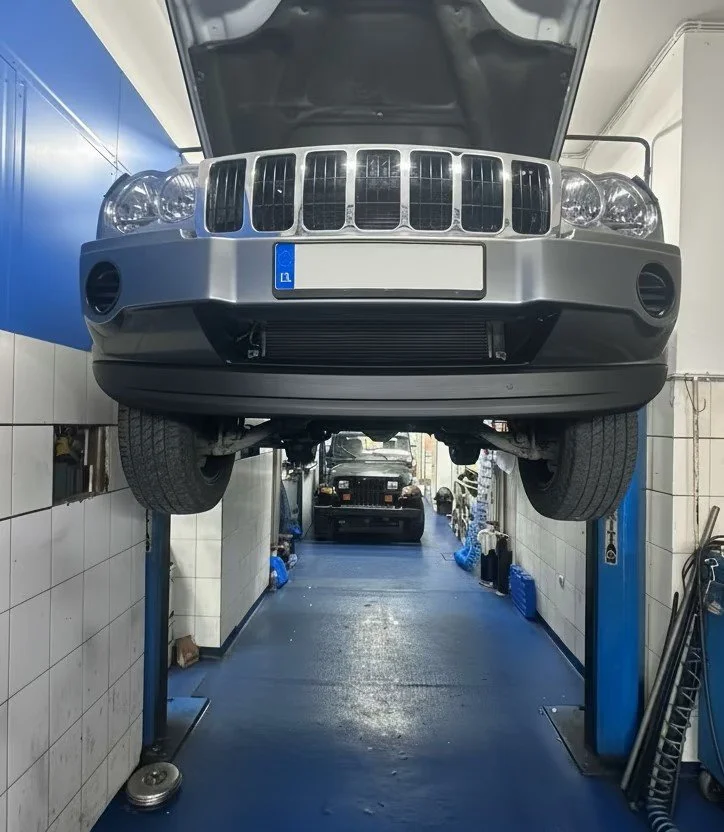 Front view of a silver car lifted on a hydraulic lift inside an automotive repair shop, with the underside and front wheels visible.