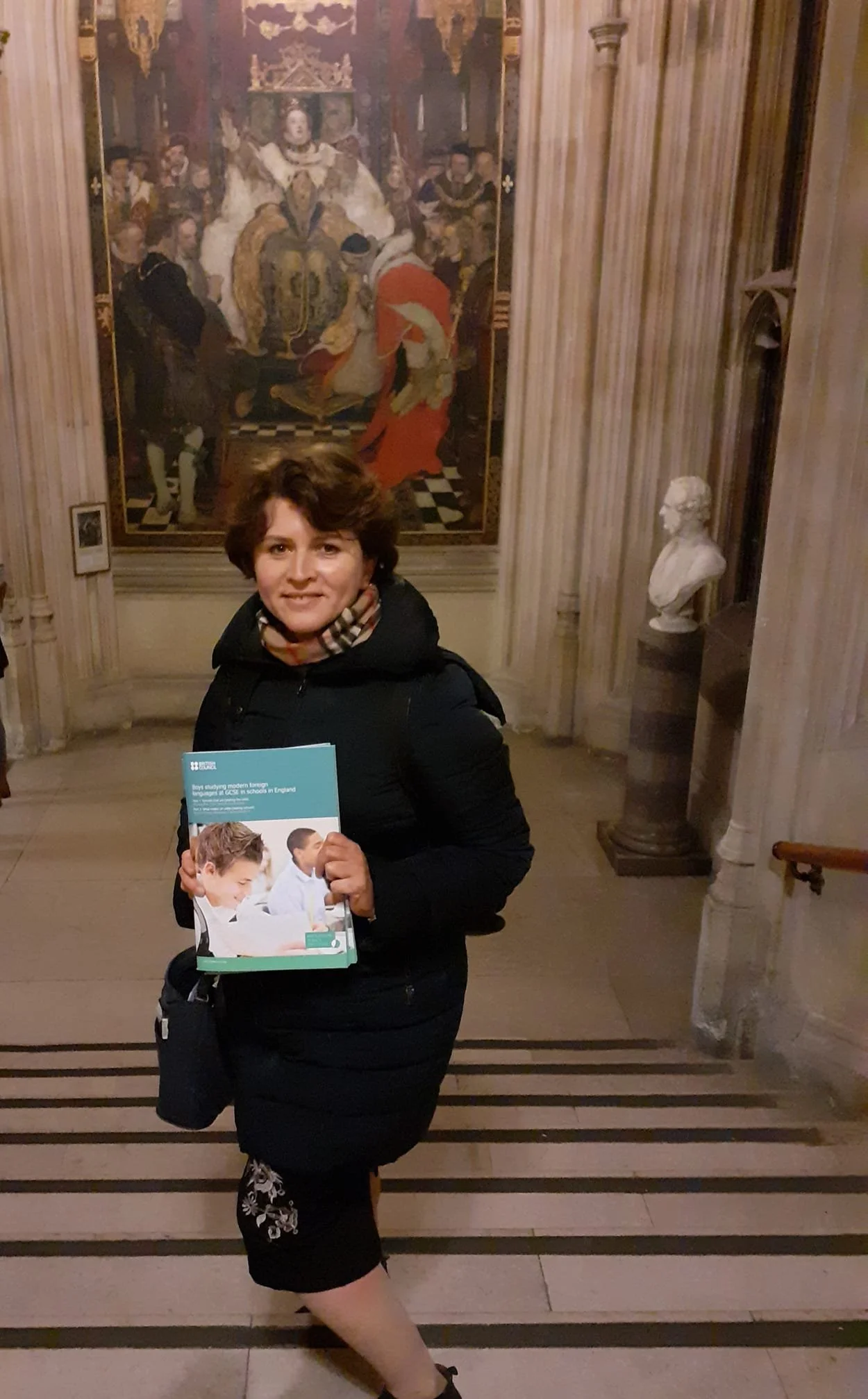 A woman holding a booklet stands in front of a painting in a historic building with marble columns and a bust sculpture.
