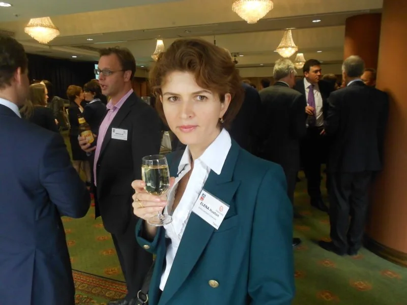 Woman with short brown hair wearing a teal blazer and white shirt holding a glass of champagne at a professional networking event with other attendees in the background.