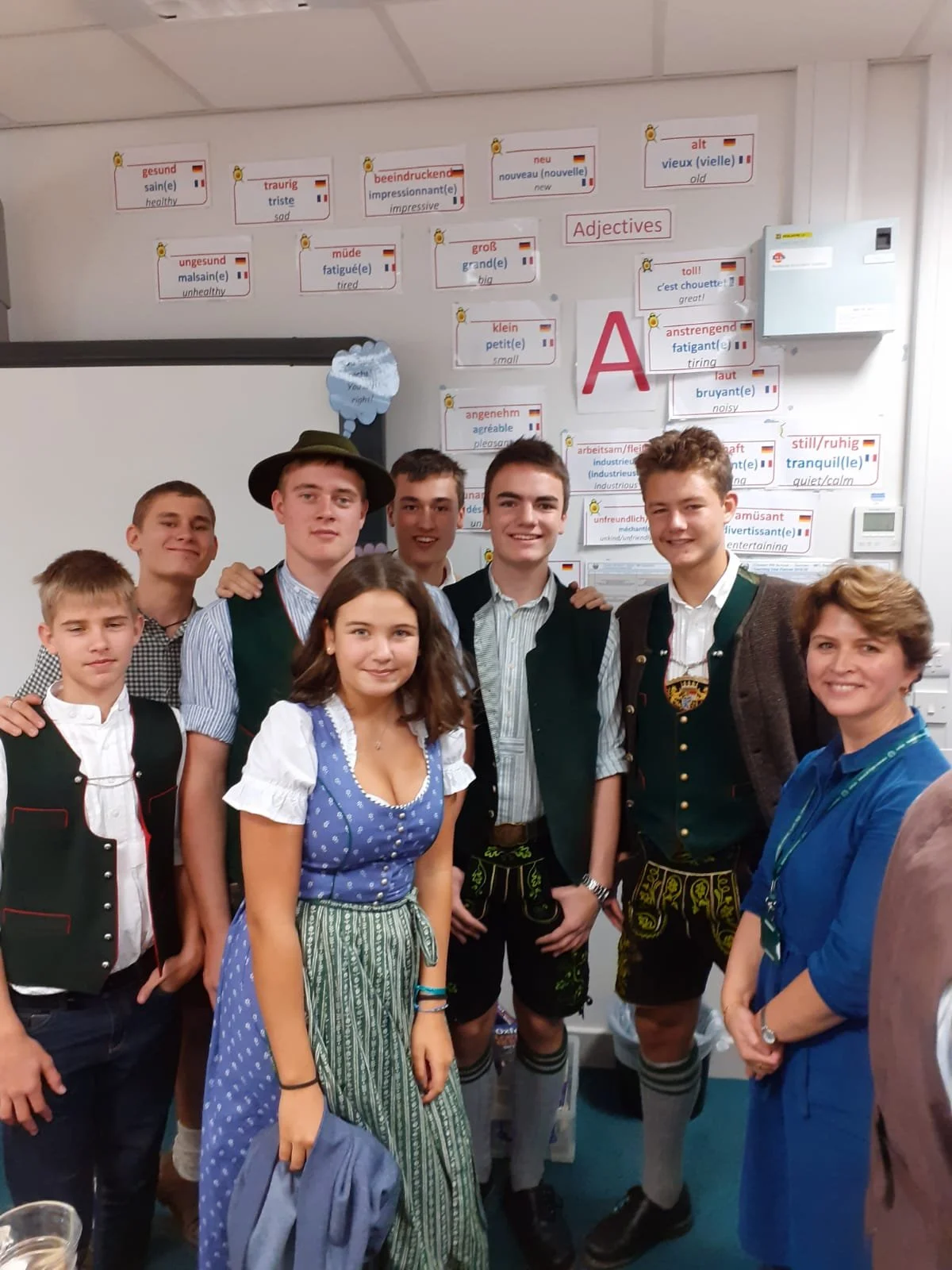 Group of young people in traditional Bavarian clothing posing in a classroom with educational posters about German adjectives on the wall.
