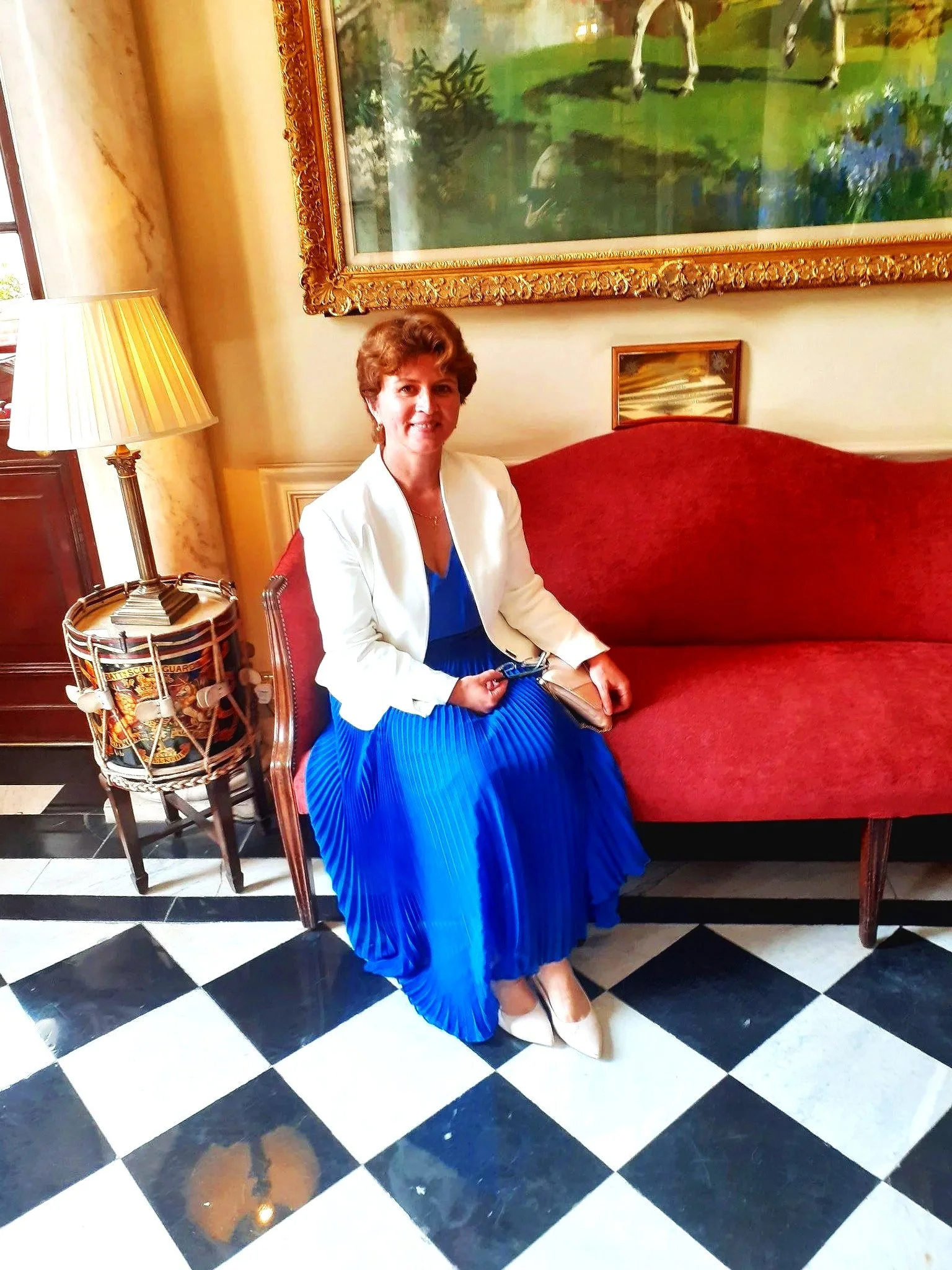 Woman in a blue dress and white jacket sitting on a red vintage sofa in a decorated room with a framed painting hanging above.