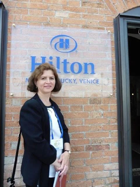 A woman standing outside a Hilton hotel in Louisville, Kentucky, Venice area, with a brick wall and Hilton sign behind her.
