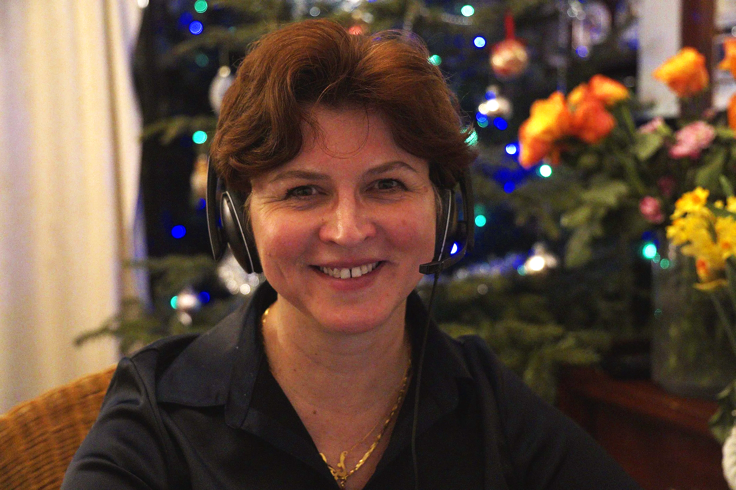 A smiling woman with short brown hair, wearing a black shirt and a gold necklace, sitting indoors in front of a decorated Christmas tree with colorful lights and ornaments, while wearing a headset.