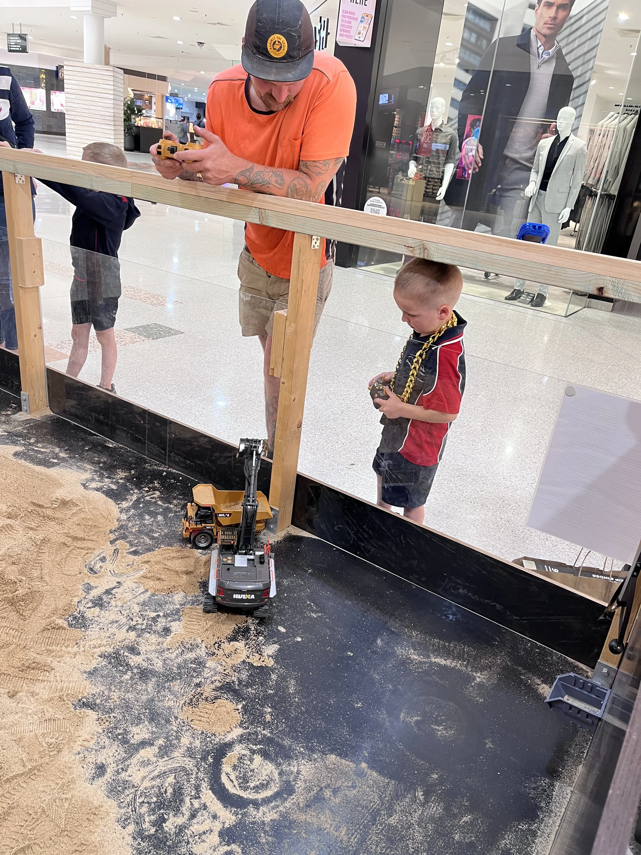A man and two children are playing in an indoor sandbox at a shopping mall. The man, wearing an orange shirt and a cap, is holding a remote control device. The children are standing inside the sandbox, one with a toy excavator and the other with a toy dump truck. There is a toy excavator in the sand and a wooden barrier separating the sandbox from the shopping area.