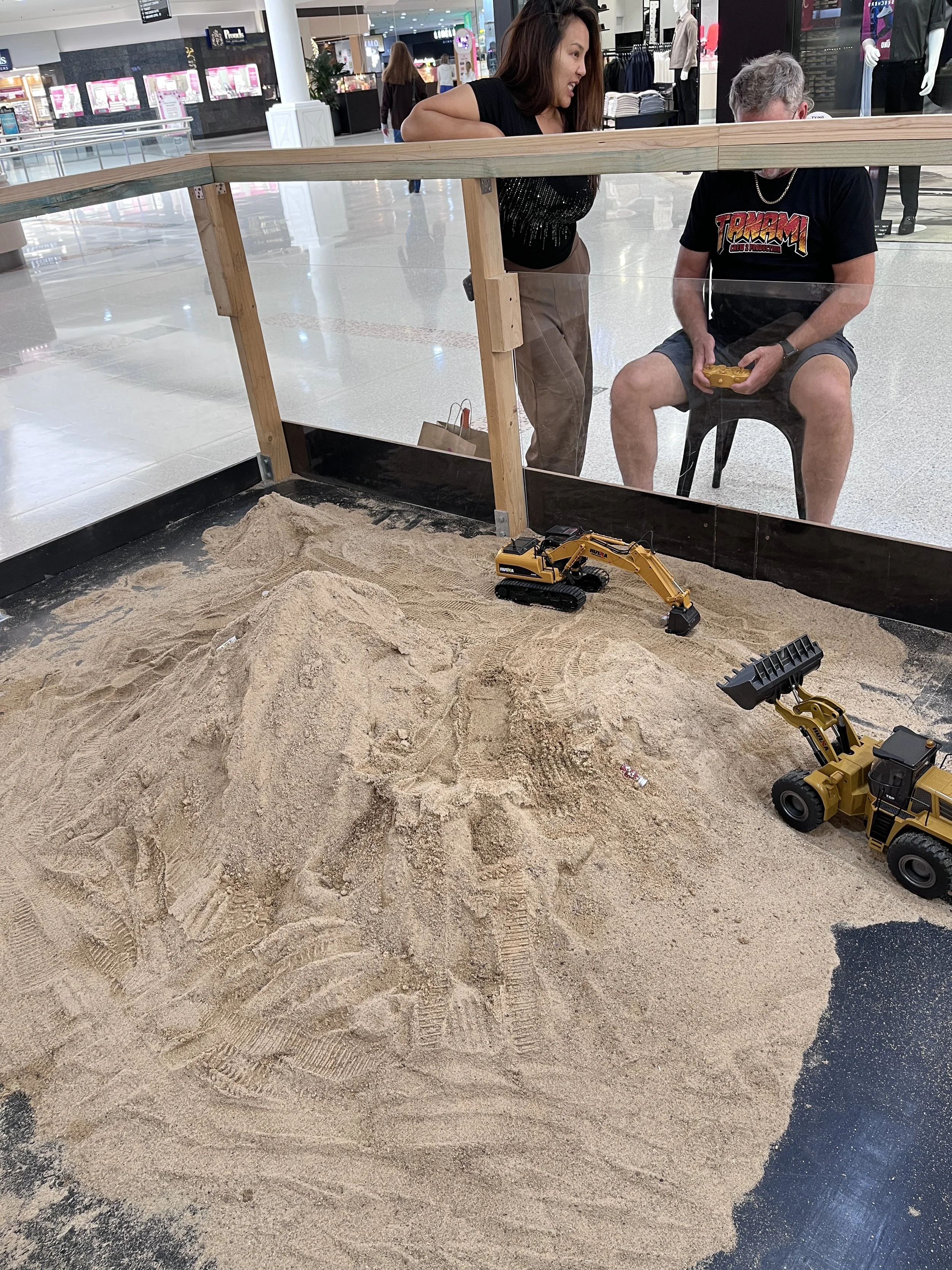 People interacting over a sandbox on a shopping mall floor with toy construction vehicles, sand, and a wood barrier.