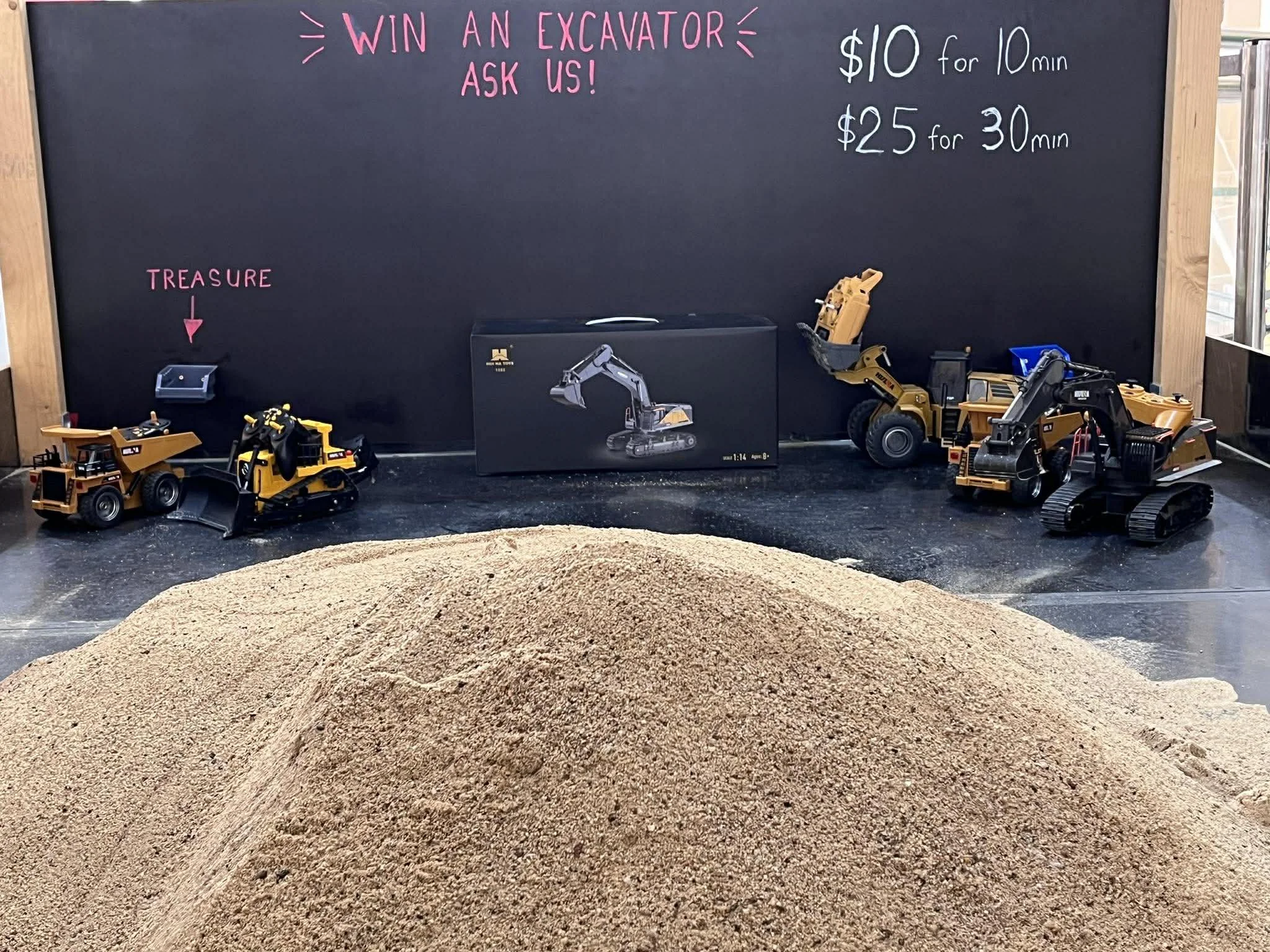 A display board advertising a contest to win a toy excavator, with toy excavators, a box, and a pile of dirt or sand in front of it. The board has prices listed for 10 minutes and 30 minutes sessions.