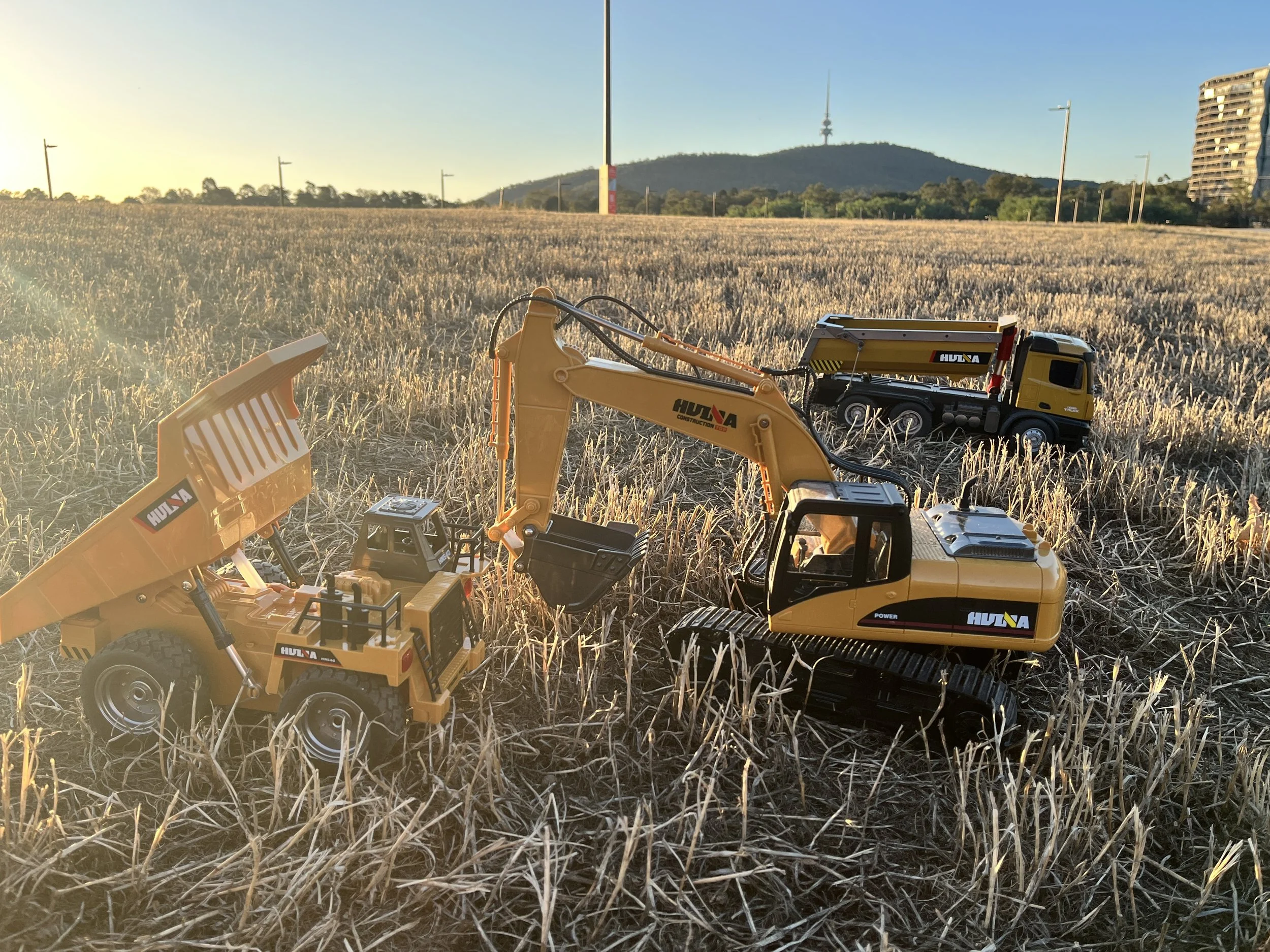 Toy construction vehicles, including an excavator, dump truck, and front loader, are placed in a field with dry grass during sunset, with mountains and a telecommunications tower in the background.
Canberra & NSW