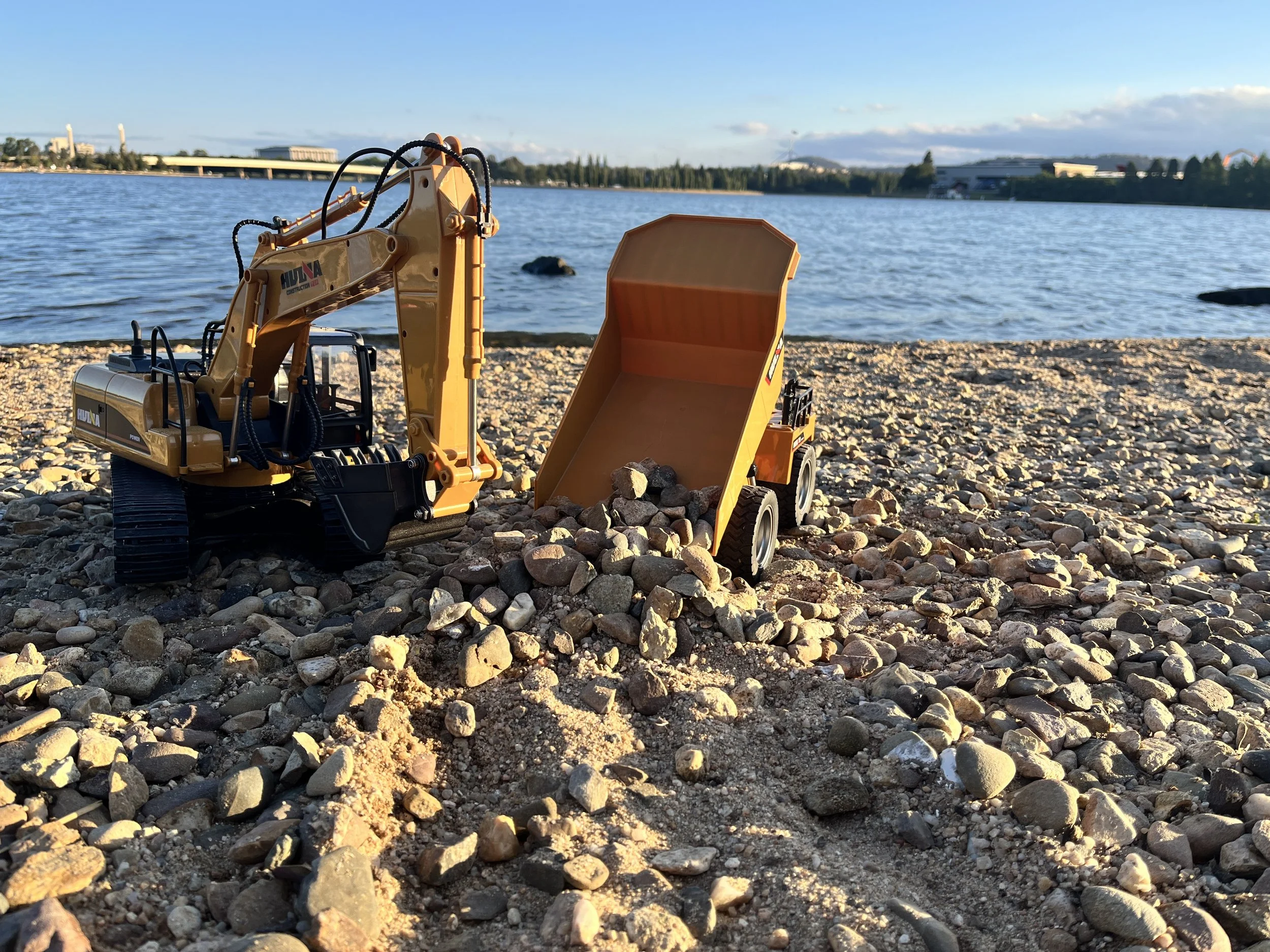 Toy construction excavator and dump truck on pebbly beach with water and buildings in the background
Canberra & NSW