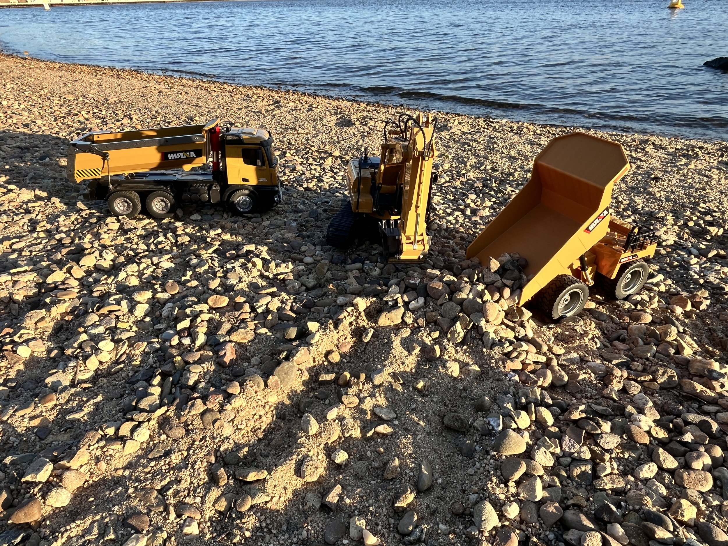 Three toy construction vehicles, a dump truck, an excavator, and a loader, placed on pebbled beach near water.
Canberra & NSW