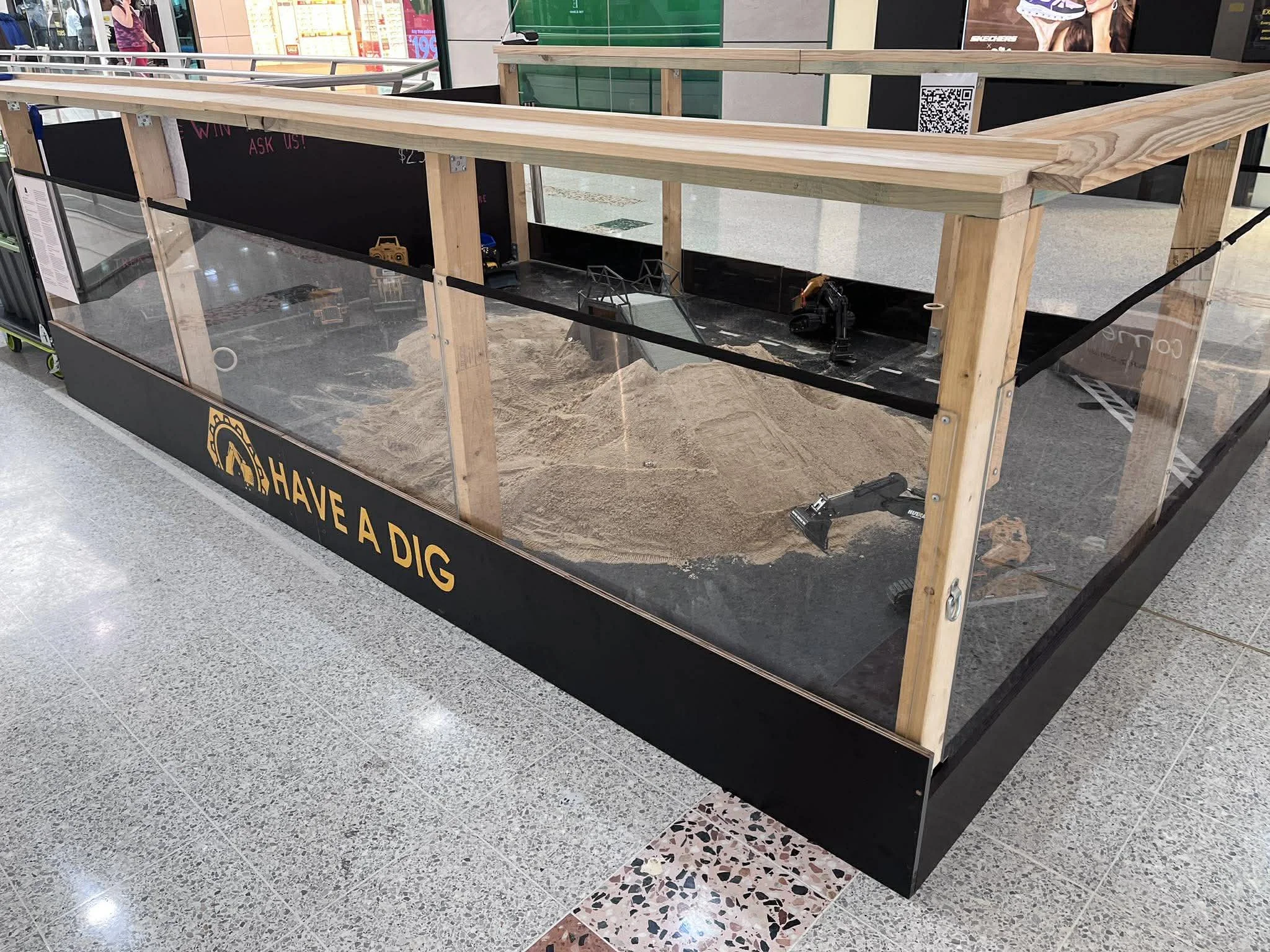Empty wooden frame with glass panels in a shopping mall, with a manual drill on the floor nearby and a black sign at the bottom that says 'HAVE A DIG'.