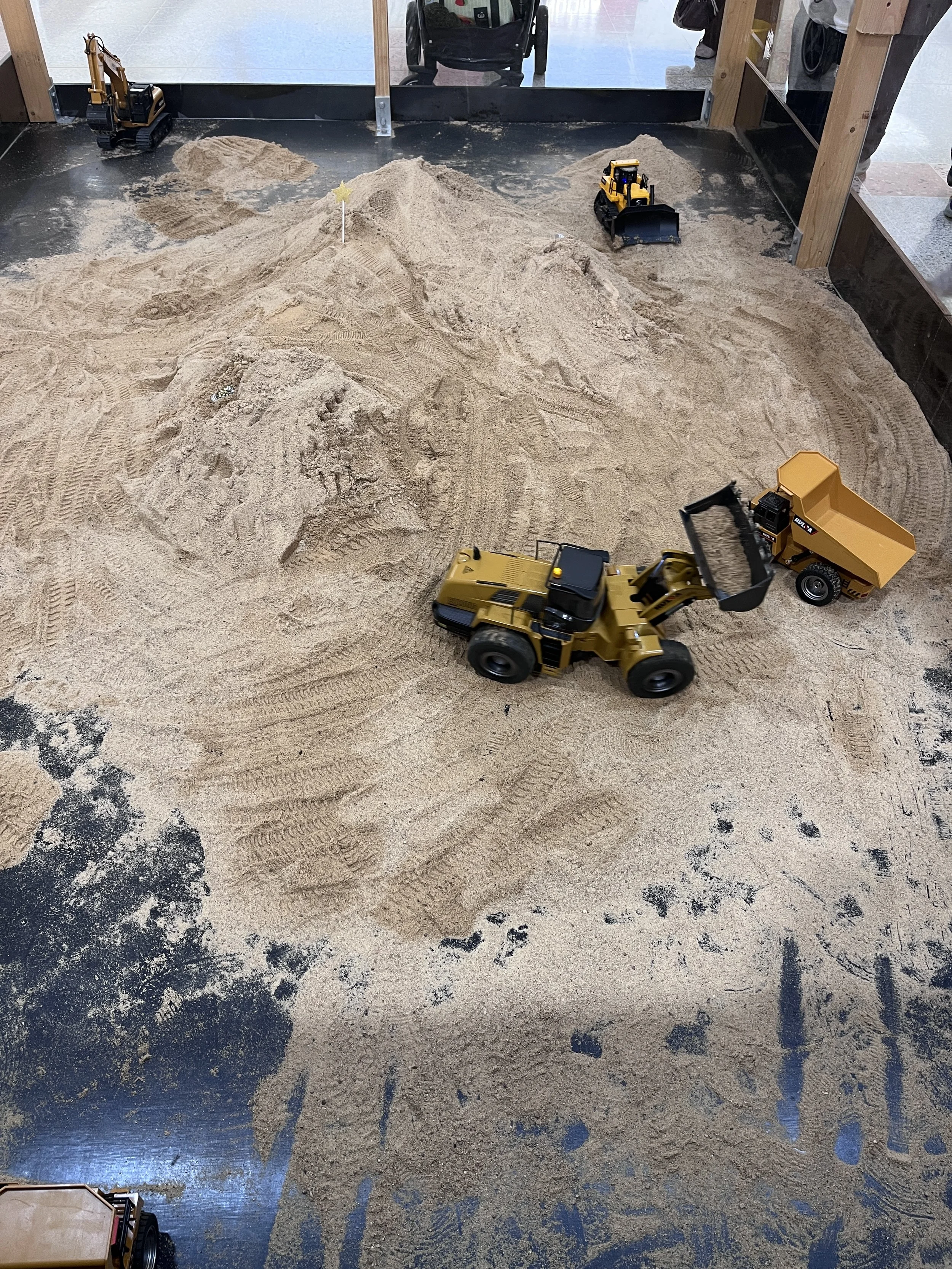 Indoor sandbox with toy construction vehicles, including a yellow bulldozer, a yellow dump truck, and a small yellow excavator. Sand is piled in the center, with children and adults partially visible around the edges.