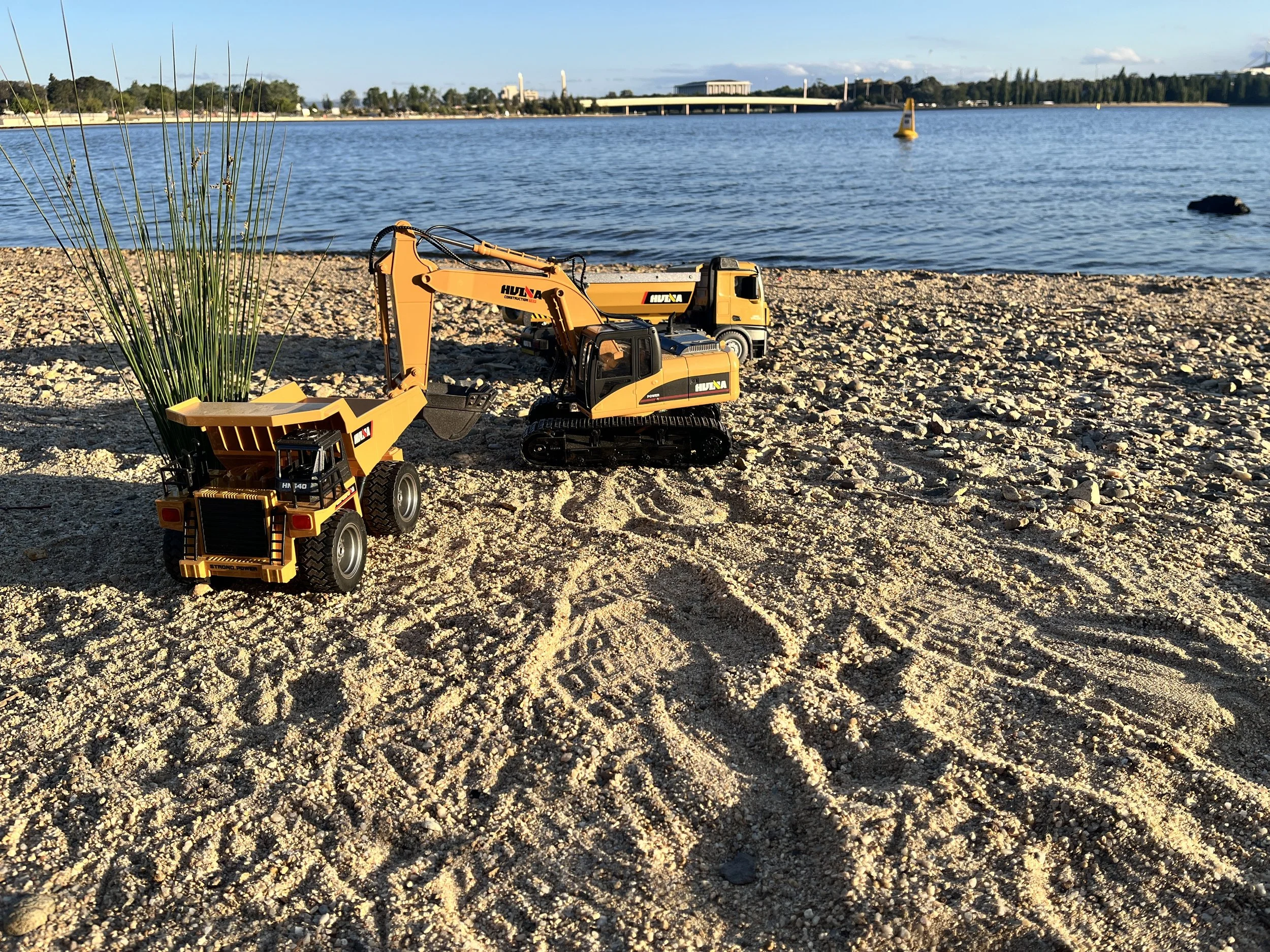 Toy construction vehicles on sandy beach with water and sailboats in the background.