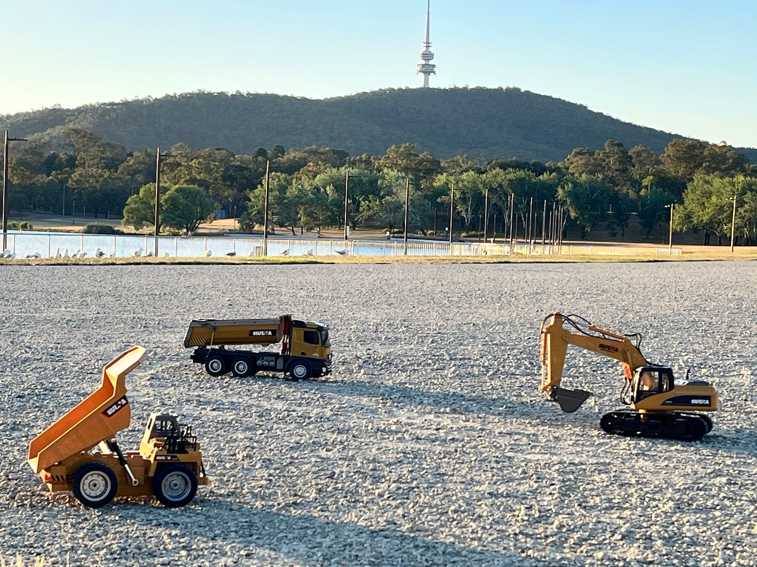 Construction equipment toys on a gravel surface with a hill and communication tower in the background.