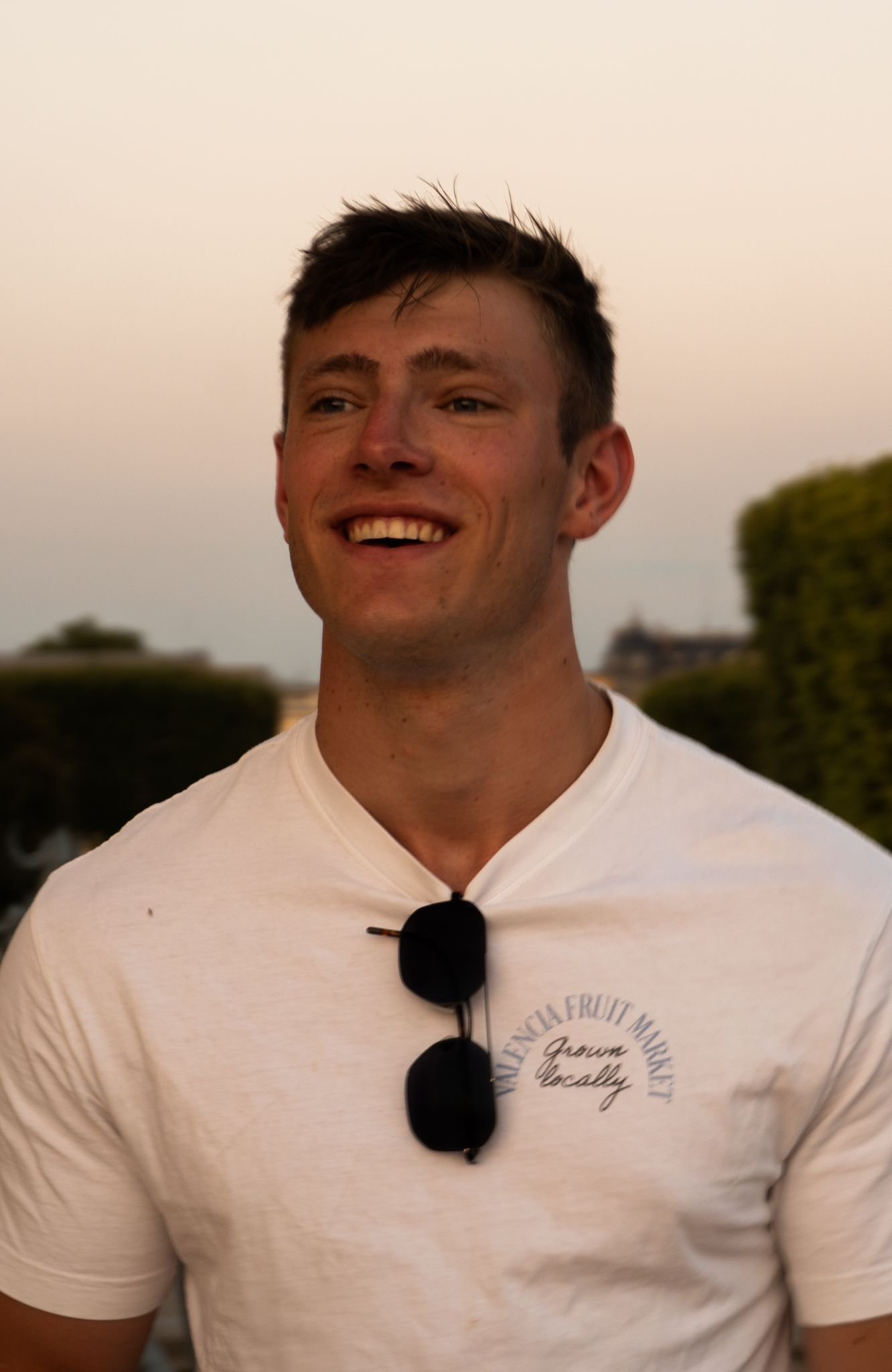 Een lachende jonge man met kort, donker haar, zittend buiten tijdens de schemering. Hij draagt een wit T-shirt met tekst en een paar zwarte zonnebrillen hangen aan de hals van zijn shirt.