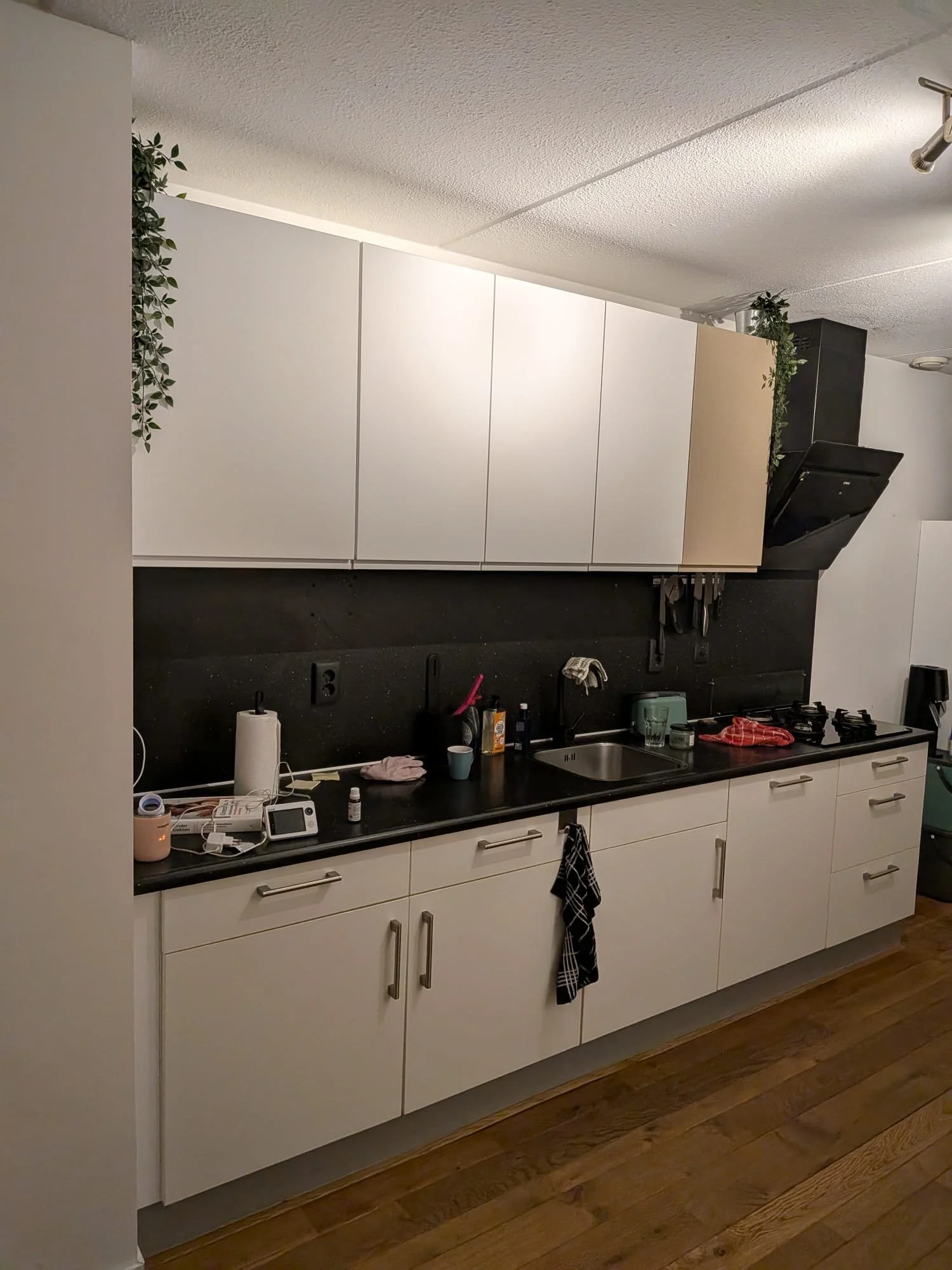 Kleine, moderne keuken met witte kasten, een zwart werkblad, een gootsteen, enkele huishoudelijke spullen en een houten vloer.