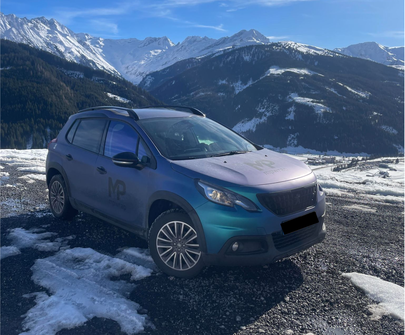 Een blauwe eilandauto met logo op de zijkant, geparkeerd op een bergpas in de sneeuw met bergen op de achtergrond.