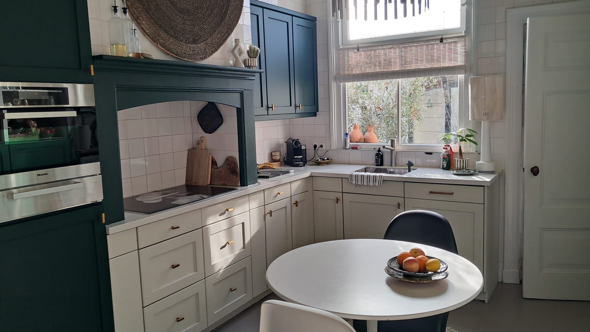 Moderne keuken met blauwe en witte kasten, ronde witte eettafel met een schaal fruit, grote ramen met verduistergordijn en planten, keukenapparatuur en decoratie op het aanrecht.