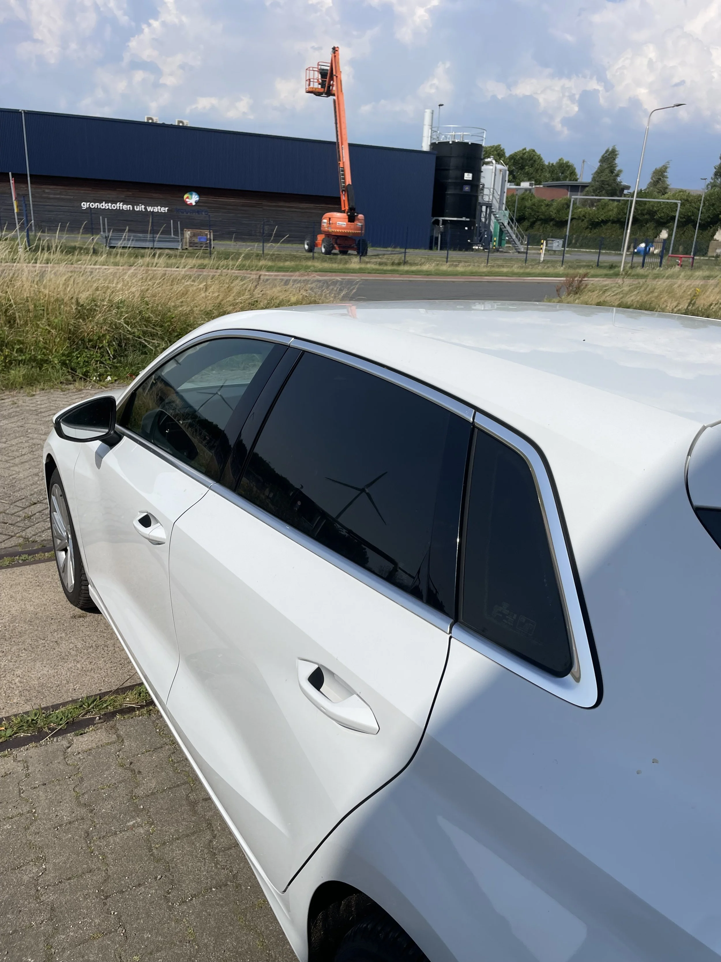 Wit hatchback auto geparkeerd op een parkeerplaats, met een bouwmachine en een industrieel gebouw op de achtergrond.