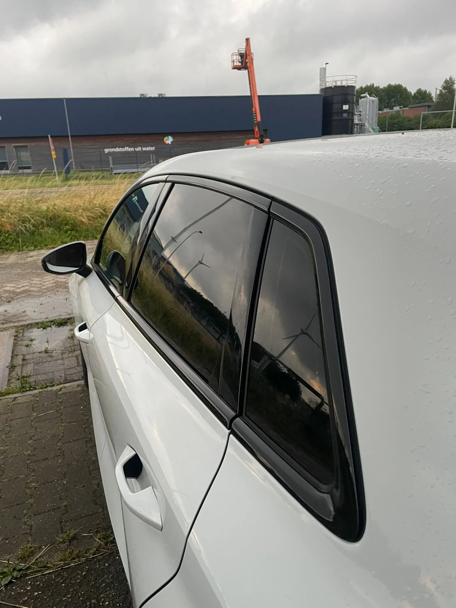 Close-up van de rechterkant van een witte auto met reflecterende ramen, parkeert op een oprit bij een bedrijf dat watergrundstoffen produceert. Het weer is bewolkt met regendruppels op de auto.