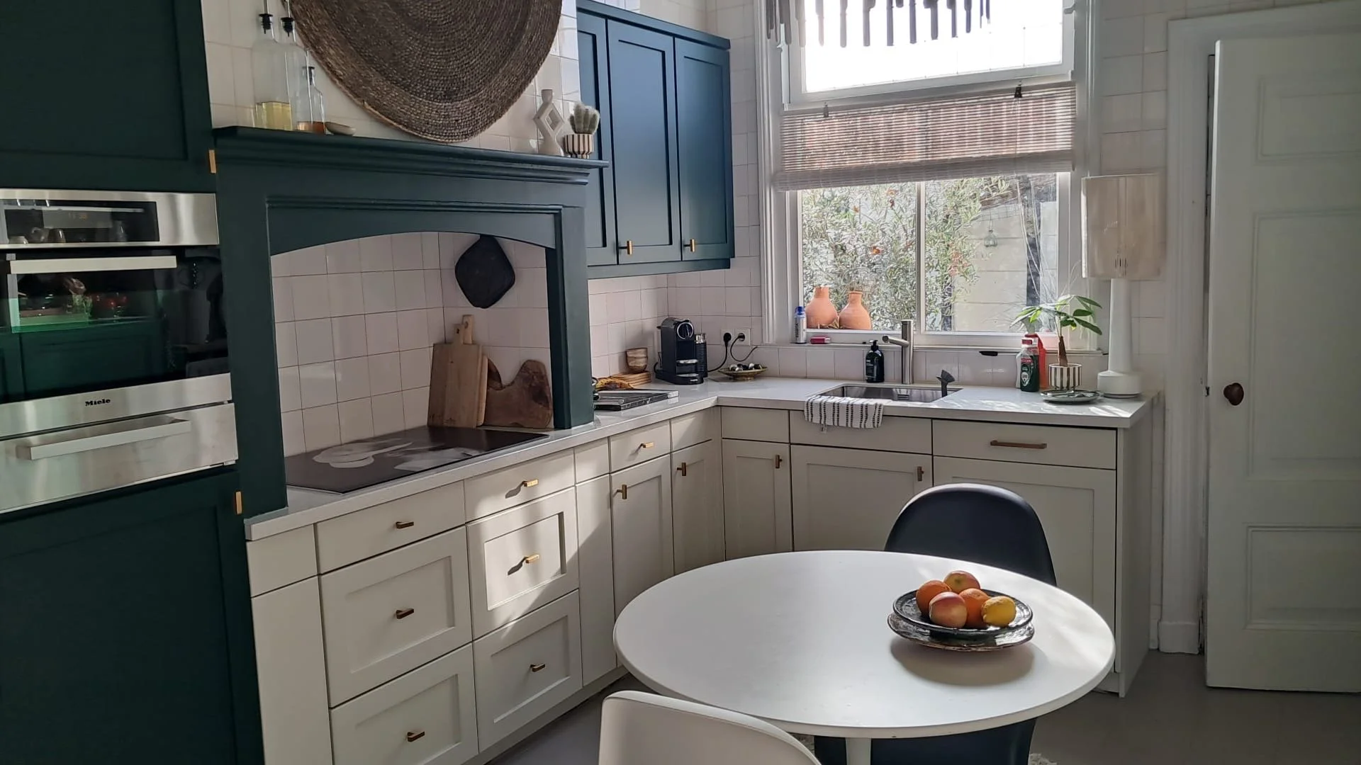 Een keuken met witte en blauwe kasten, een ronde witte tafel met een fruitschaal met appels en peren, een raam met jaloezieën en diverse keukenattributen op het aanrecht.