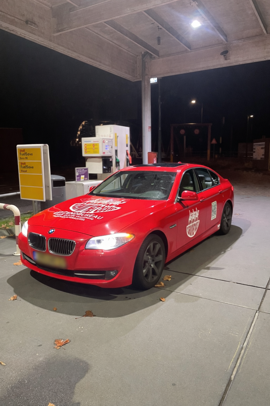 Een rode BMW-auto bij een tankstation 'Shell FuelSave' in de avondnacht, onder een overkapping met straatlichten op de achtergrond.