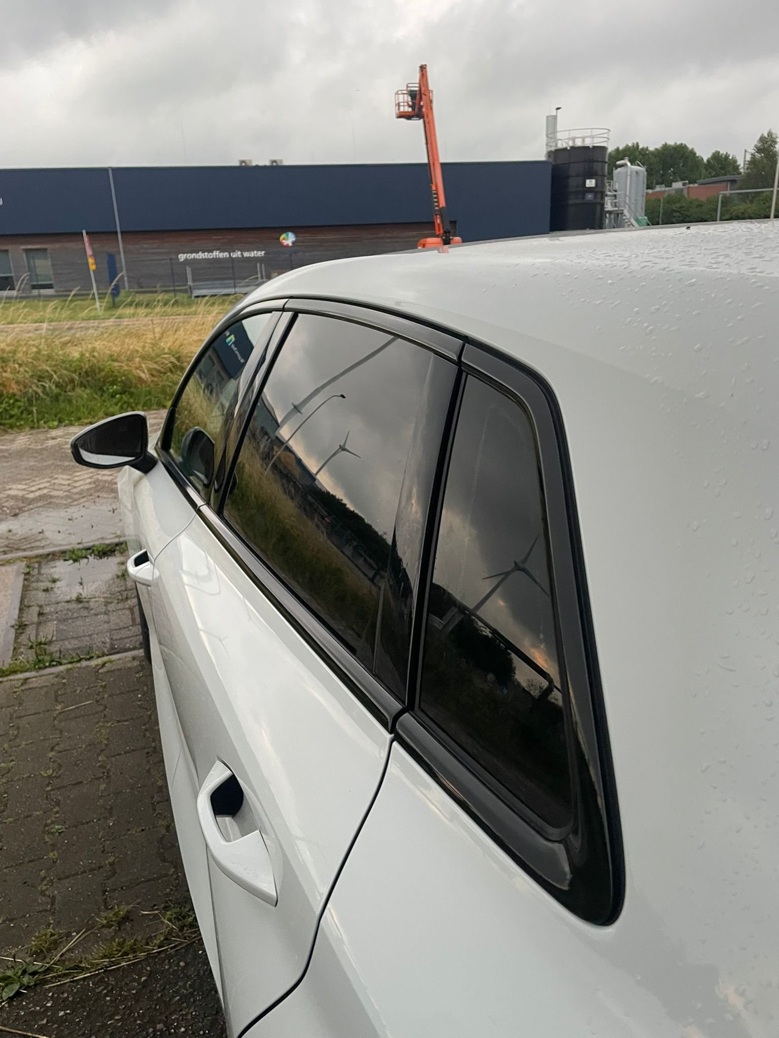Close-up van een witte auto met donkere ramen, regen druppels op het dak, geparkeerd op een stoep, met een industrieel gebouw en een rijdende hoogwerker op de achtergrond onder een bewolkte hemel.