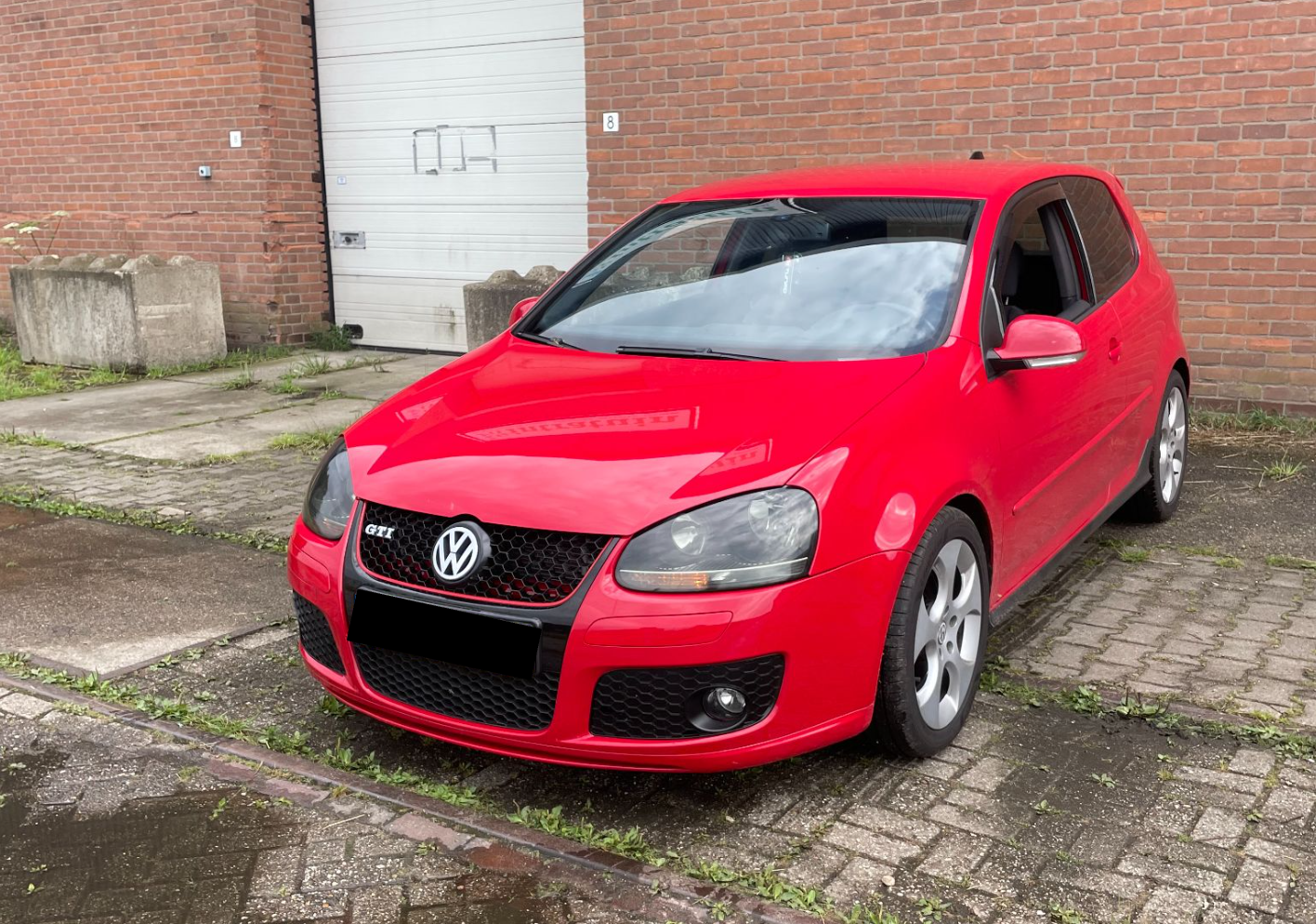 Rode Volkswagen Golf GTI geparkeerd op een oprit voor een bakstenen muur met een metalen garagedeur.