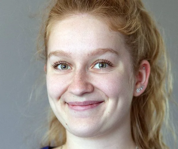 Close-up eines lachenden Mädchens mit roten Haaren, blauen Augen und mehreren Ohrpiercings vor einer grauen Wand.