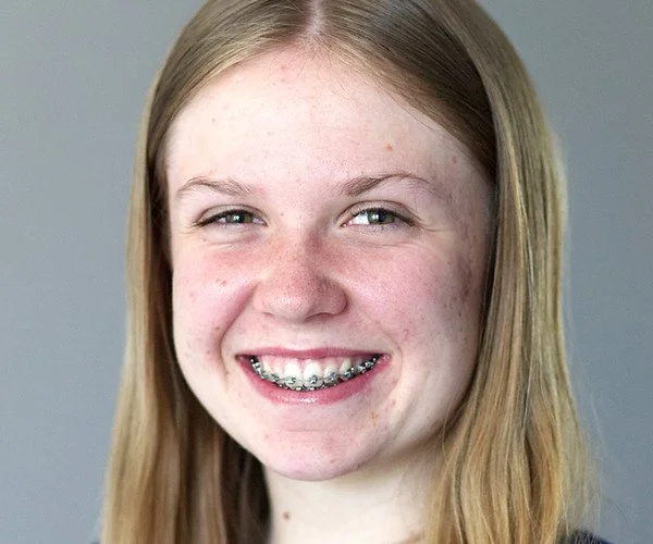 Junge Frau mit Brille und langem blonde Haar, lächelnd vor hellem Hintergrund.