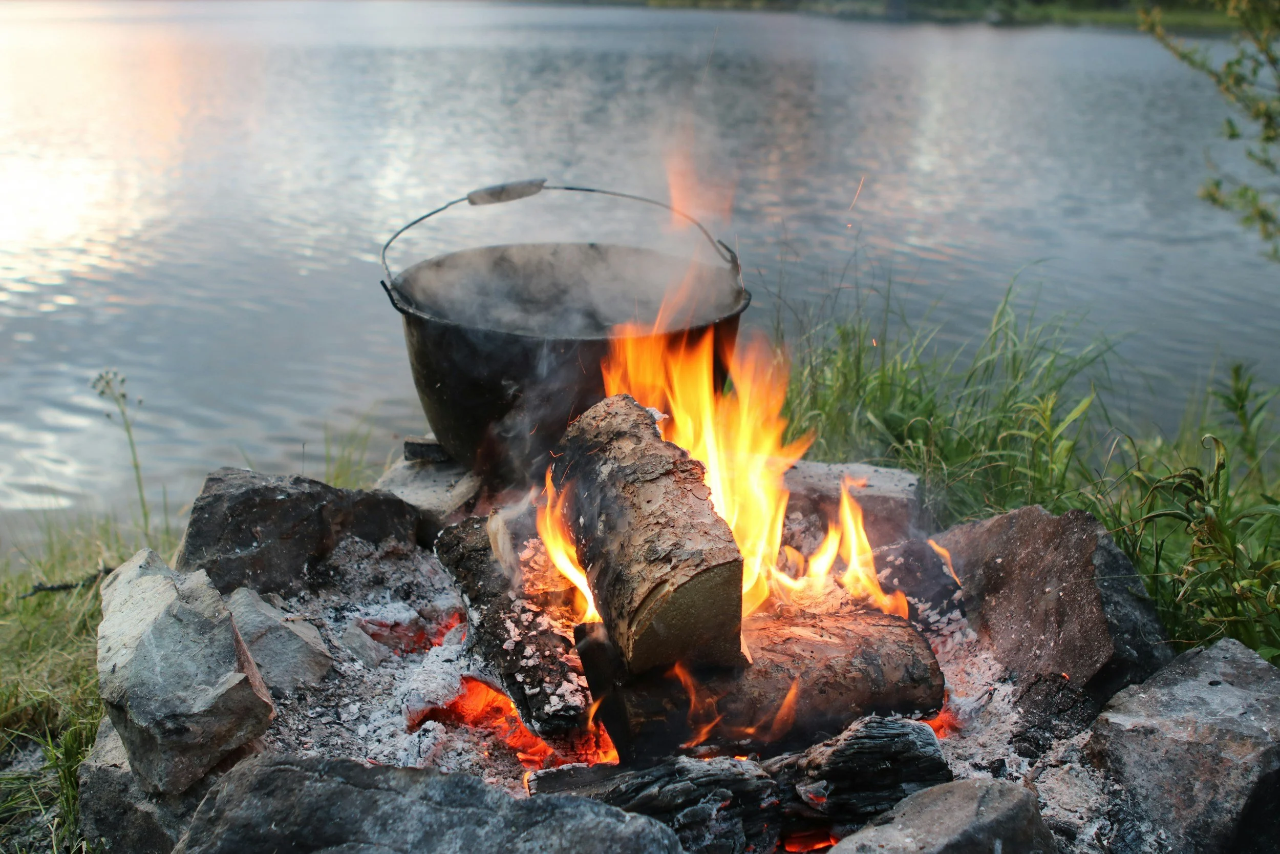 A campfire by a lakeshore with a black pot hanging over the flames, surrounded by rocks and green grass, with a body of water in the background.