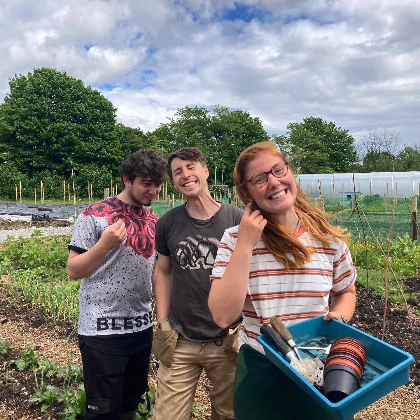 Three people standing in a garden with plants and trees in the background, smiling and posing for the photo, one woman holding a blue container with gardening tools and pots.