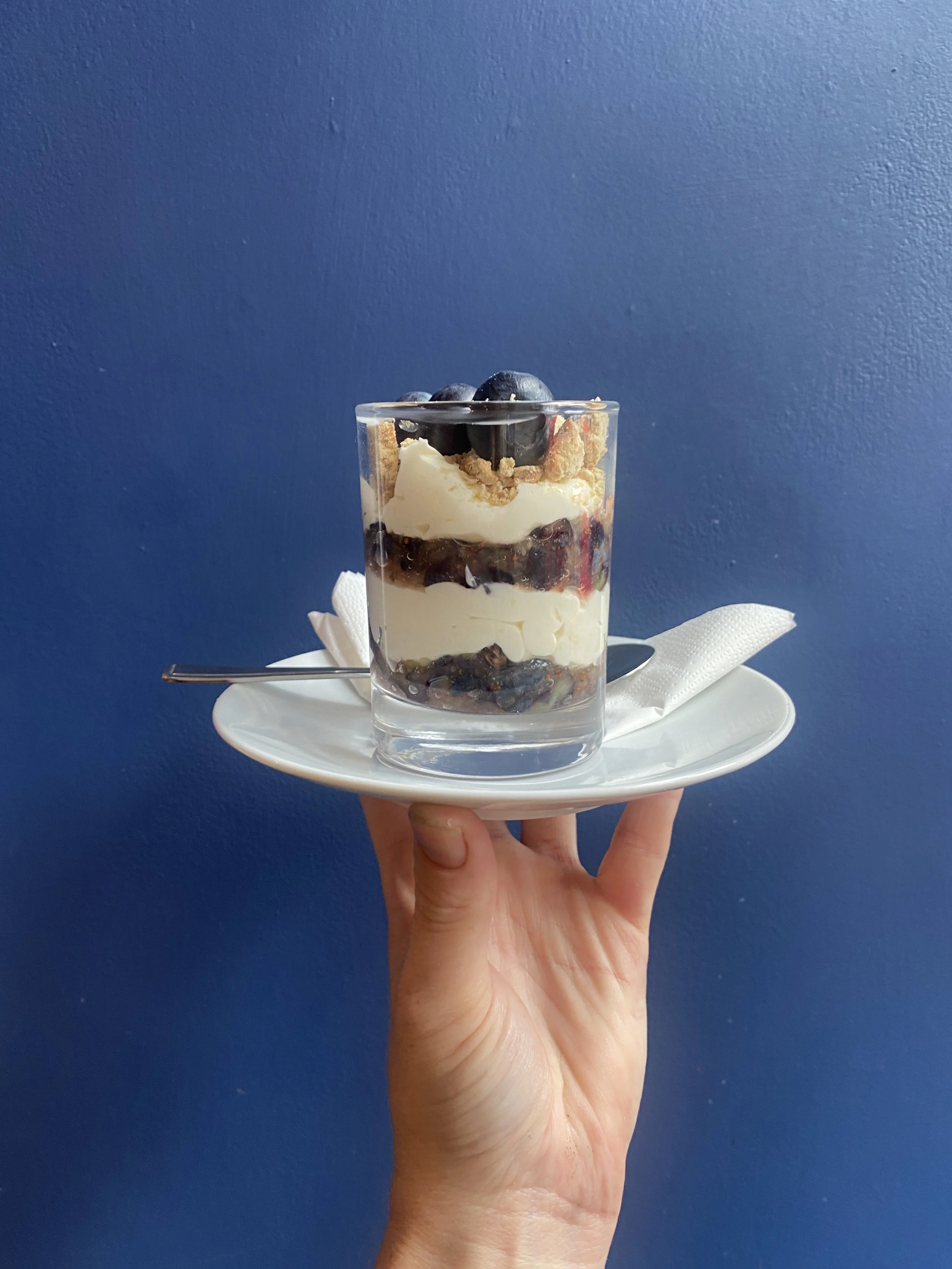 Hand holding a white plate with a small glass dessert cup filled with layered blueberries, cream, and crumble, against a blue wall background.