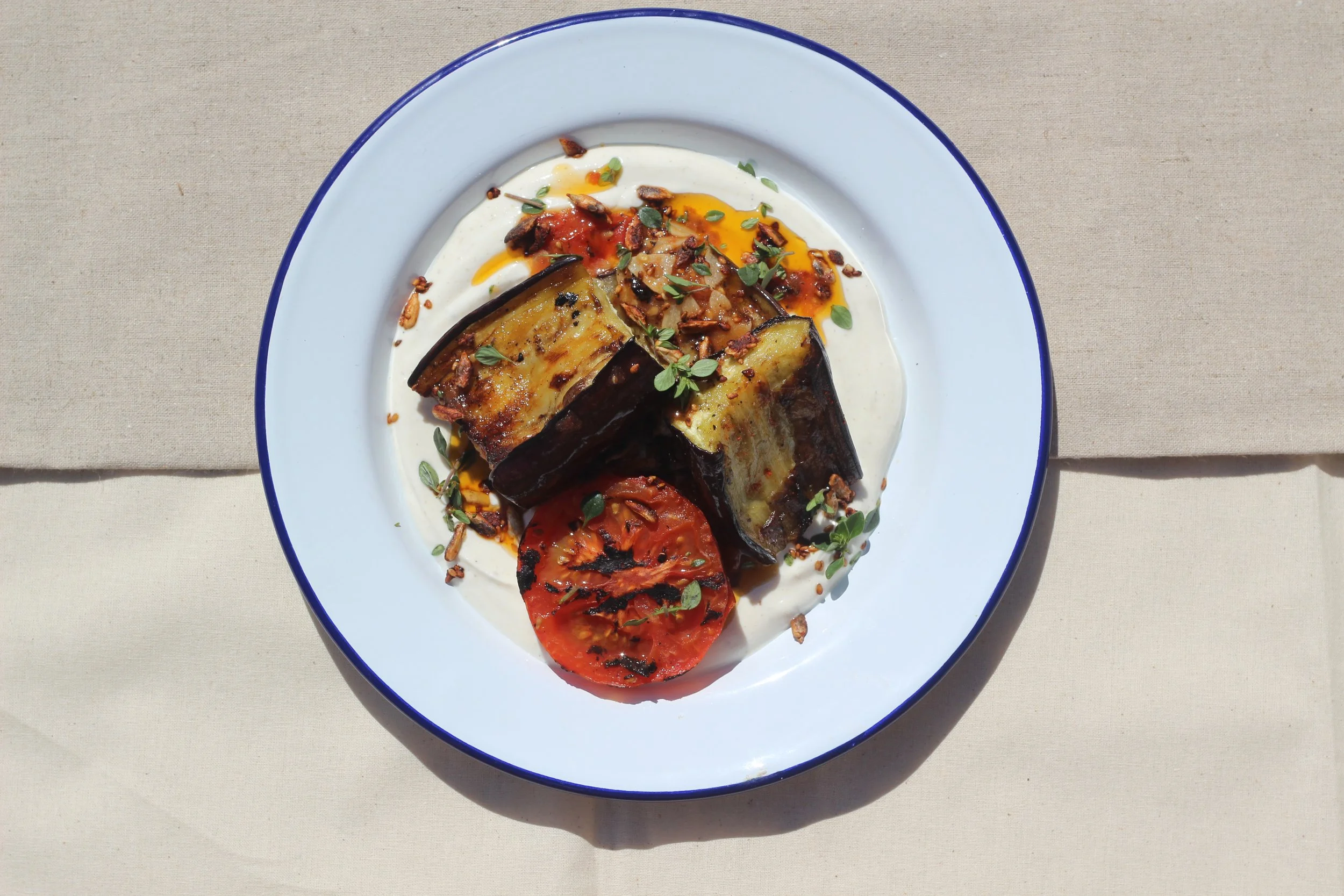 Grilled eggplant and tomato slices on a white plate with a blue rim, garnished with herbs and spices, served on a beige tablecloth.