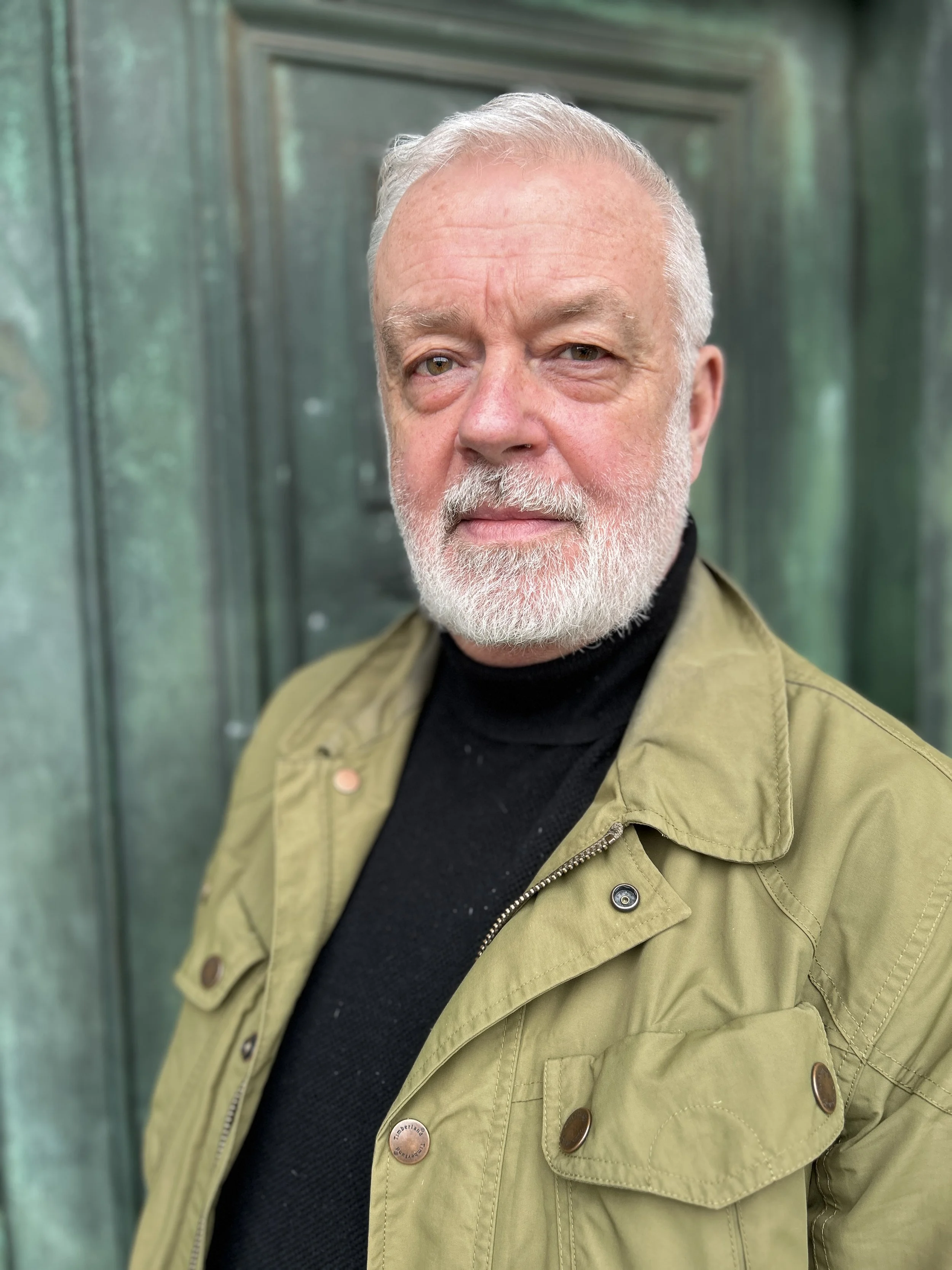 A close-up portrait of an older man with white hair and beard, wearing a black turtleneck and an olive green jacket, standing outdoors in front of a green fence.