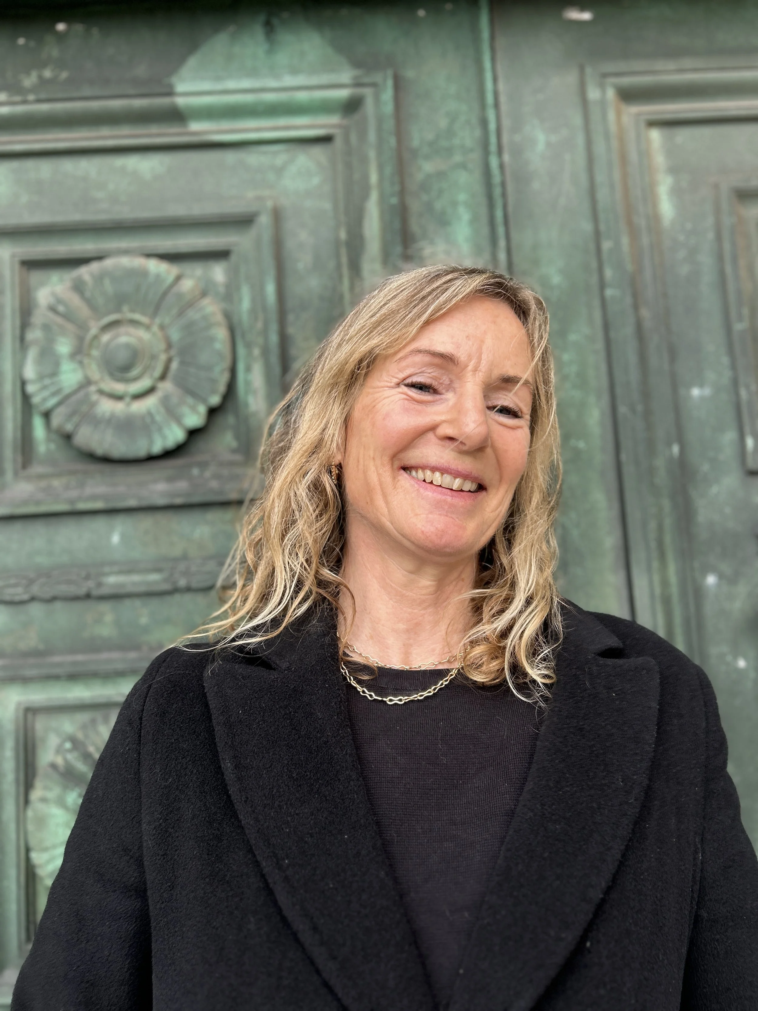 A woman with blond, wavy hair smiling, wearing a black blazer and jewelry, standing in front of a large, aged green metal door with decorative details.