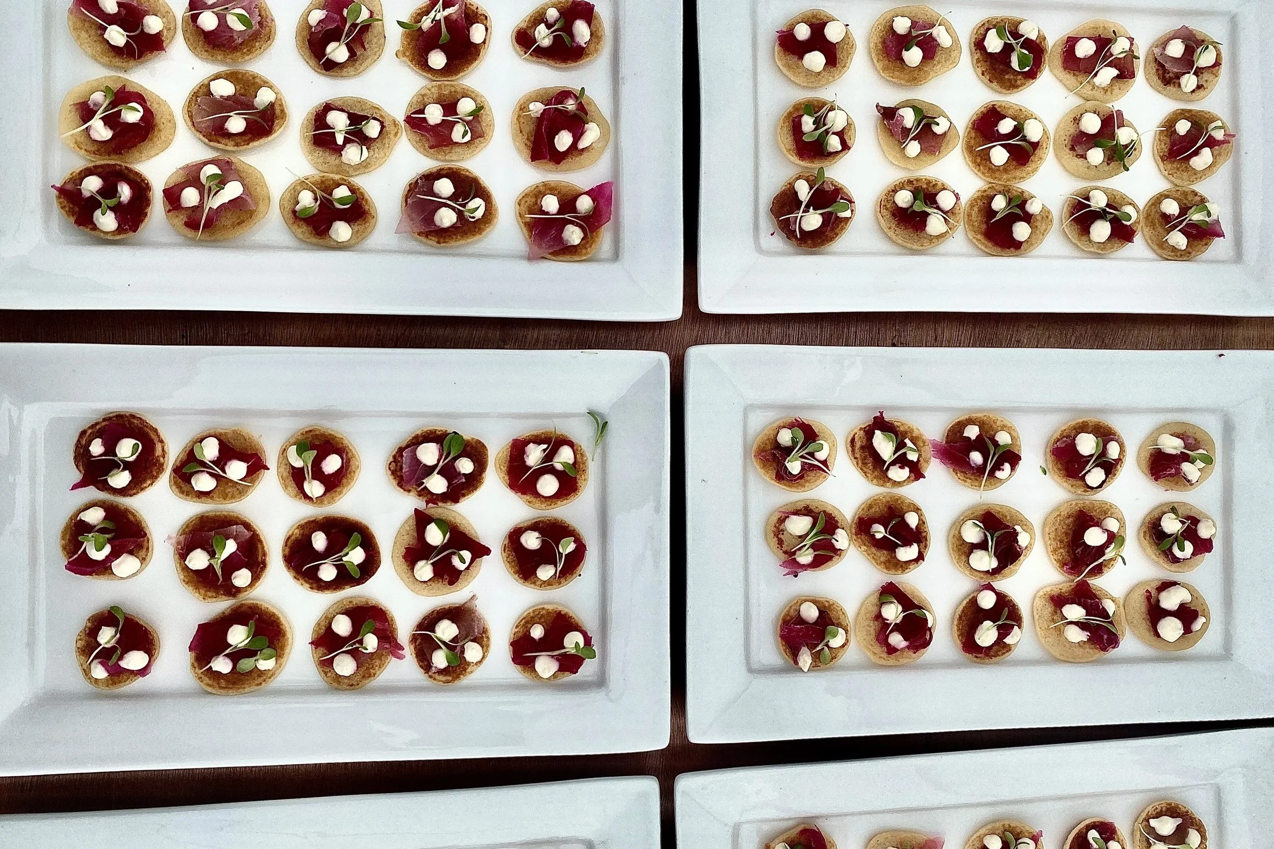 Assorted bite-sized appetizers arranged on white rectangular plates, topped with small amount of red sauce or meat, white dollops, tiny green leaves, and microgreens.