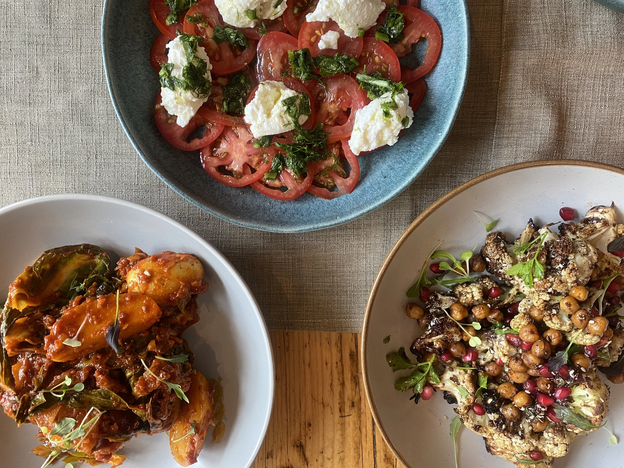 A top-down view of three dishes: a tomato and cheese salad with basil on a blue plate, a roasted vegetable dish with peppers and herbs on a white plate, and a roasted cauliflower salad with chickpeas, pomegranate seeds, and greens on a beige plate.